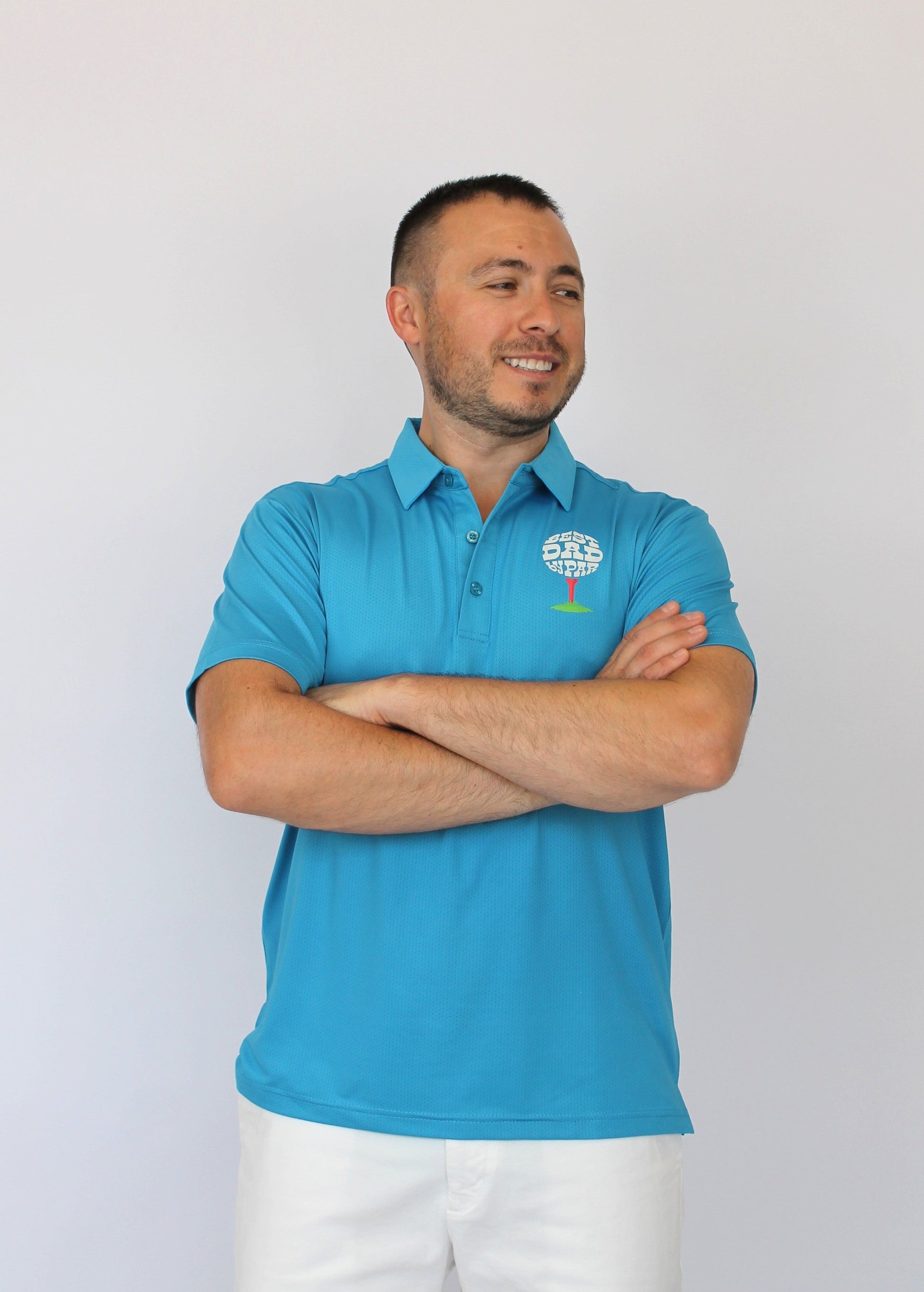 A man wearing a bright blue polo shirt with a small white and green logo on the left chest, paired with white pants, standing against a plain light gray background with his arms crossed.