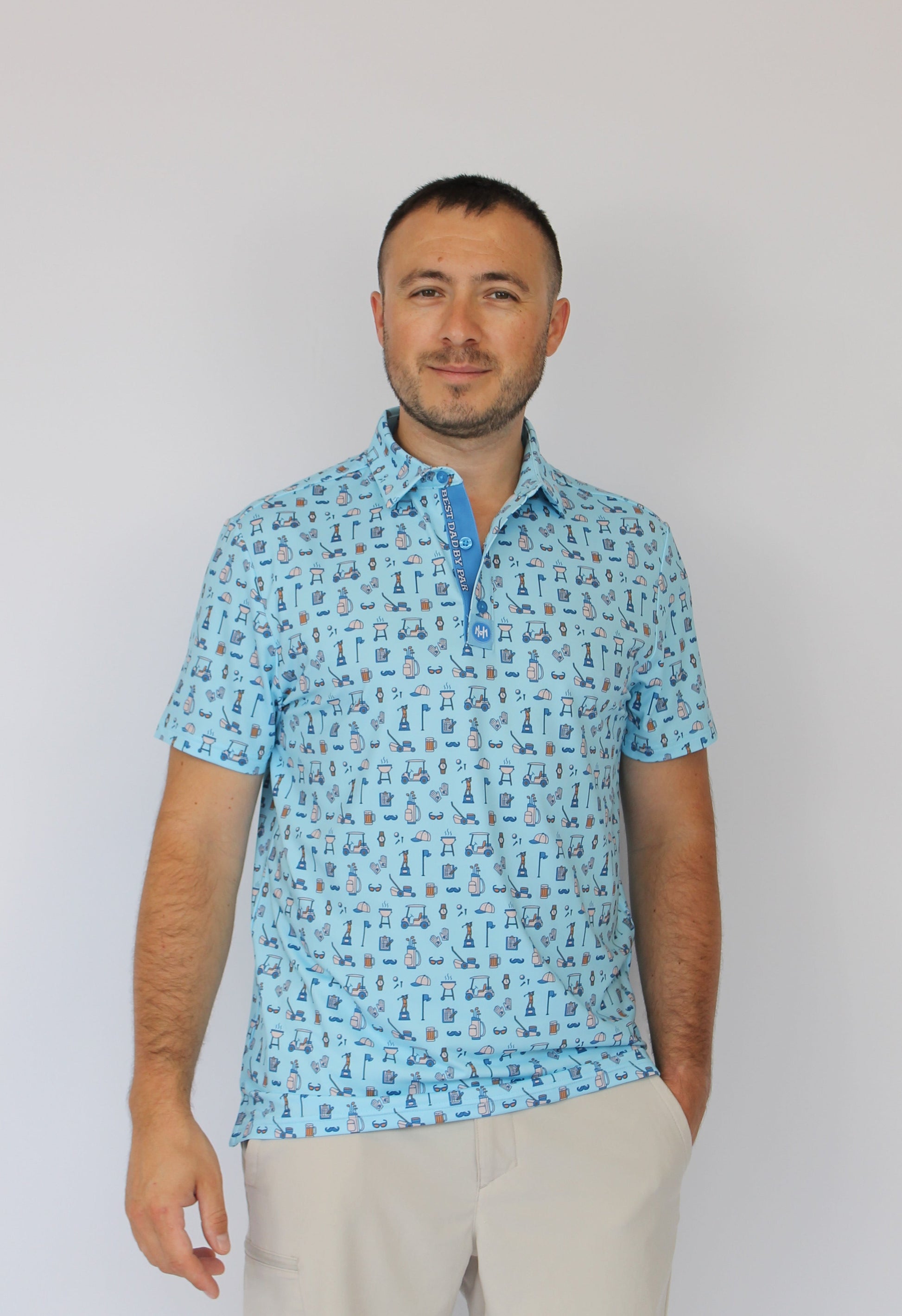 A man wearing a light blue short-sleeve polo shirt with Golf dad repeating blue pattern, paired with light beige trousers, photographed against a plain white background.