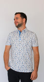A man wearing a short-sleeve polo shirt with a white base and a blue Dad-themed pattern of small golf carts, beers, golf trophy, golf flags, watch, dad grill, mustache and sunglasses, paired with dark trousers, standing against a plain light gray background.
