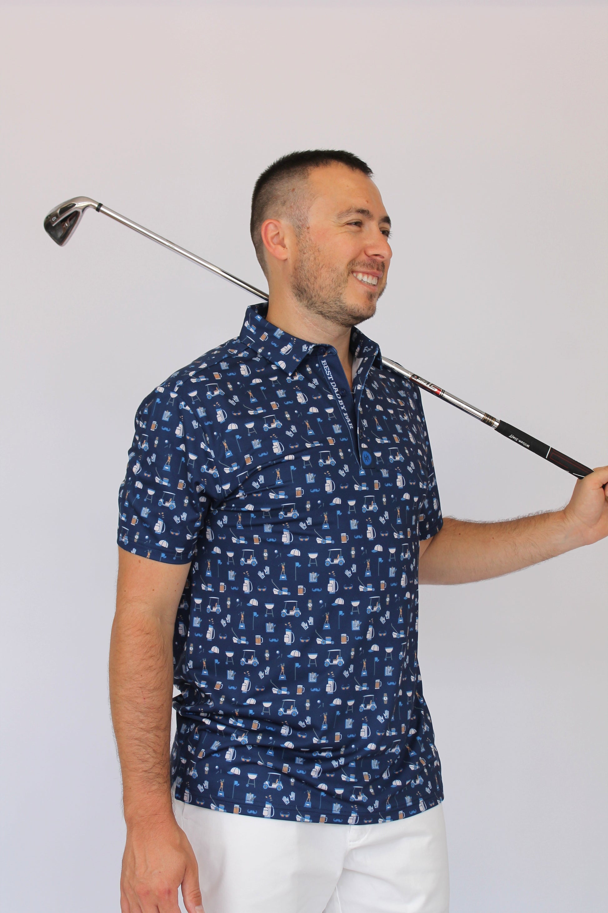 A man wearing a navy blue short-sleeve polo shirt with a white and light blue golf dad-themed pattern, holding a golf club over his shoulder against a plain light background.