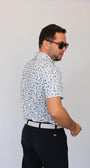 A man wearing a white short-sleeve button-up shirt with a blue Golf Dad pattern, paired with dark trousers and sunglasses, shown from a side-back angle against a plain light gray background.