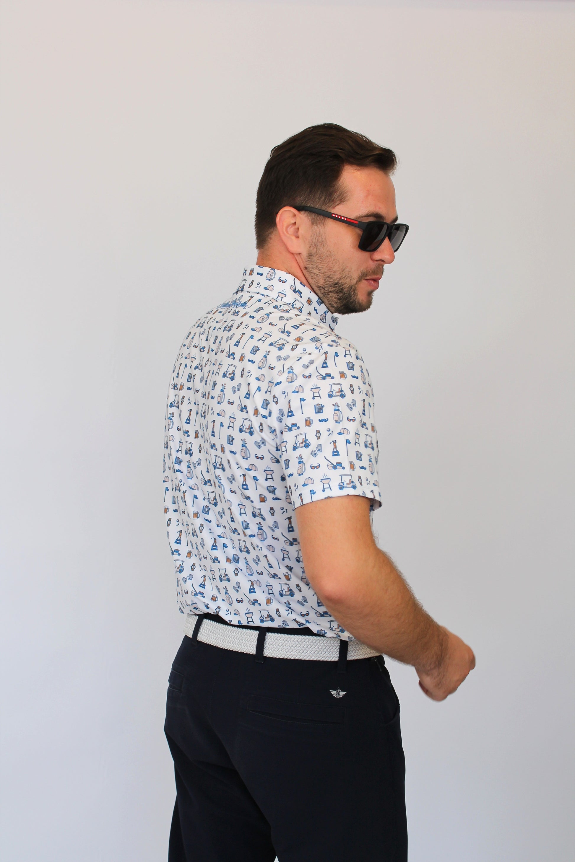 A man wearing a white short-sleeve button-up shirt with a blue Golf Dad pattern, paired with dark trousers and sunglasses, shown from a side-back angle against a plain light gray background.
