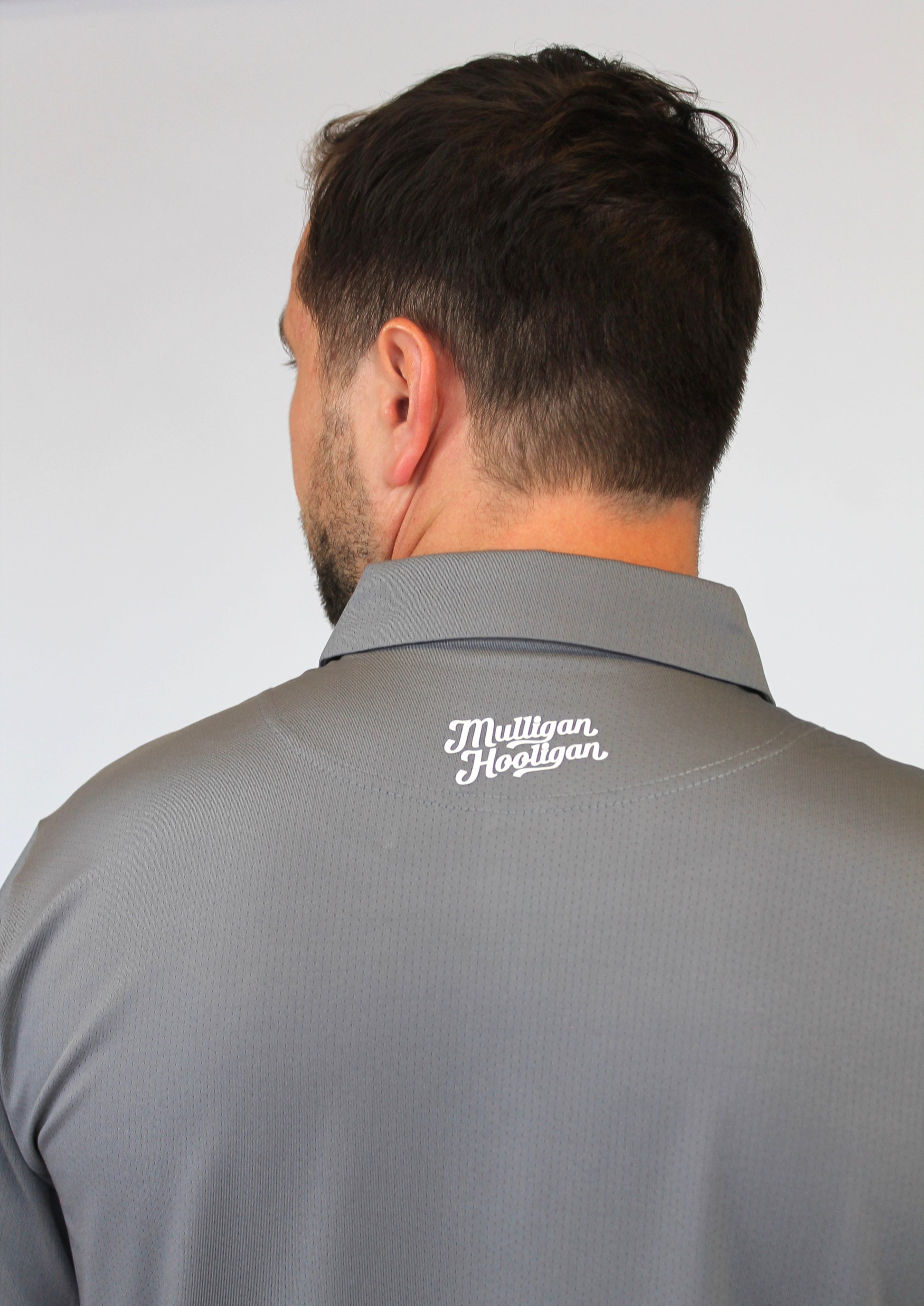 Back view of a man wearing a gray collared polo shirt with 'Mulligan Hooligan' 3D  in white script on the upper neck, against a plain light gray background.