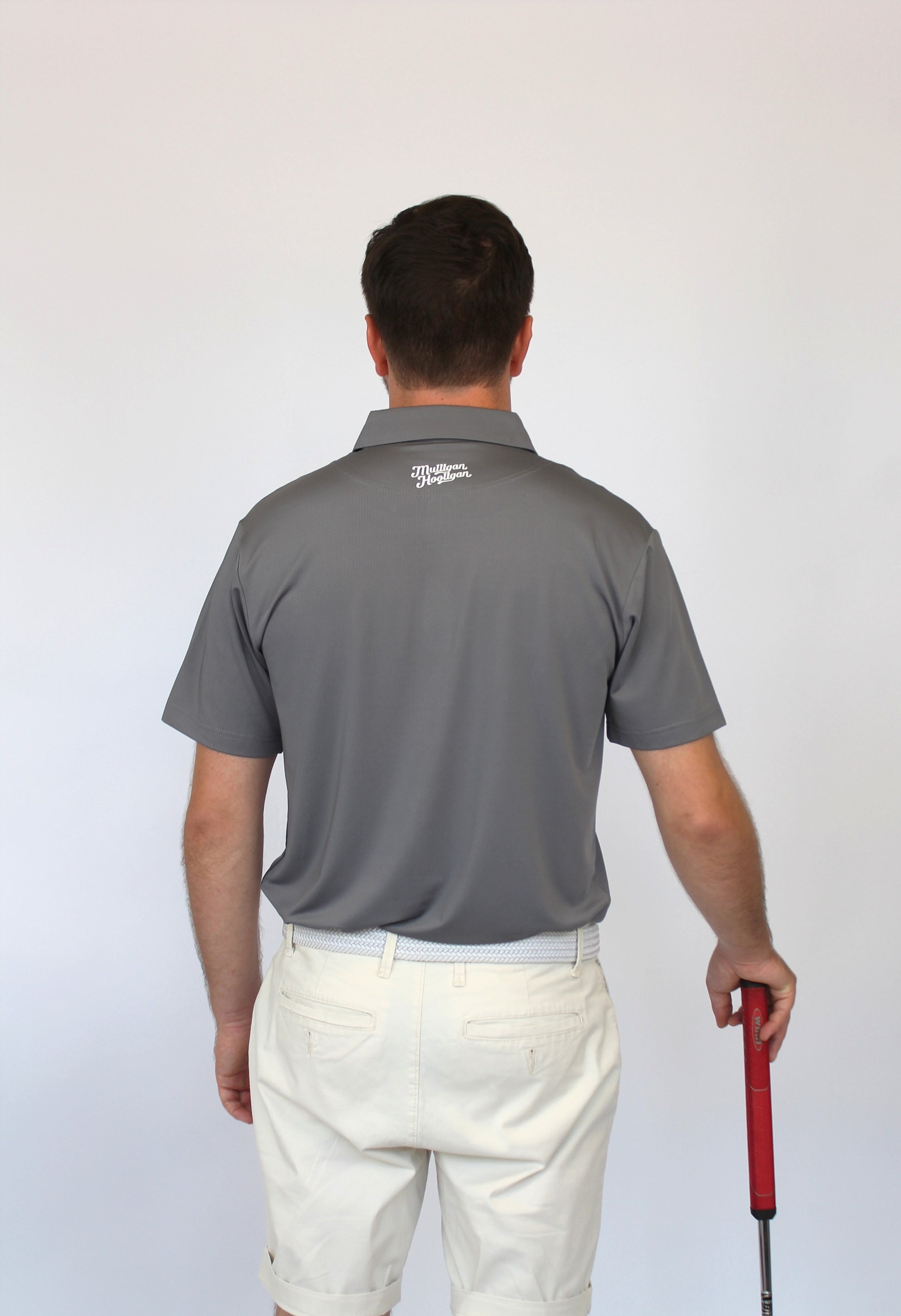 A man viewed from behind wearing a gray short-sleeve polo shirt and off-white shorts, holding a golf club with a red grip against a plain light background.