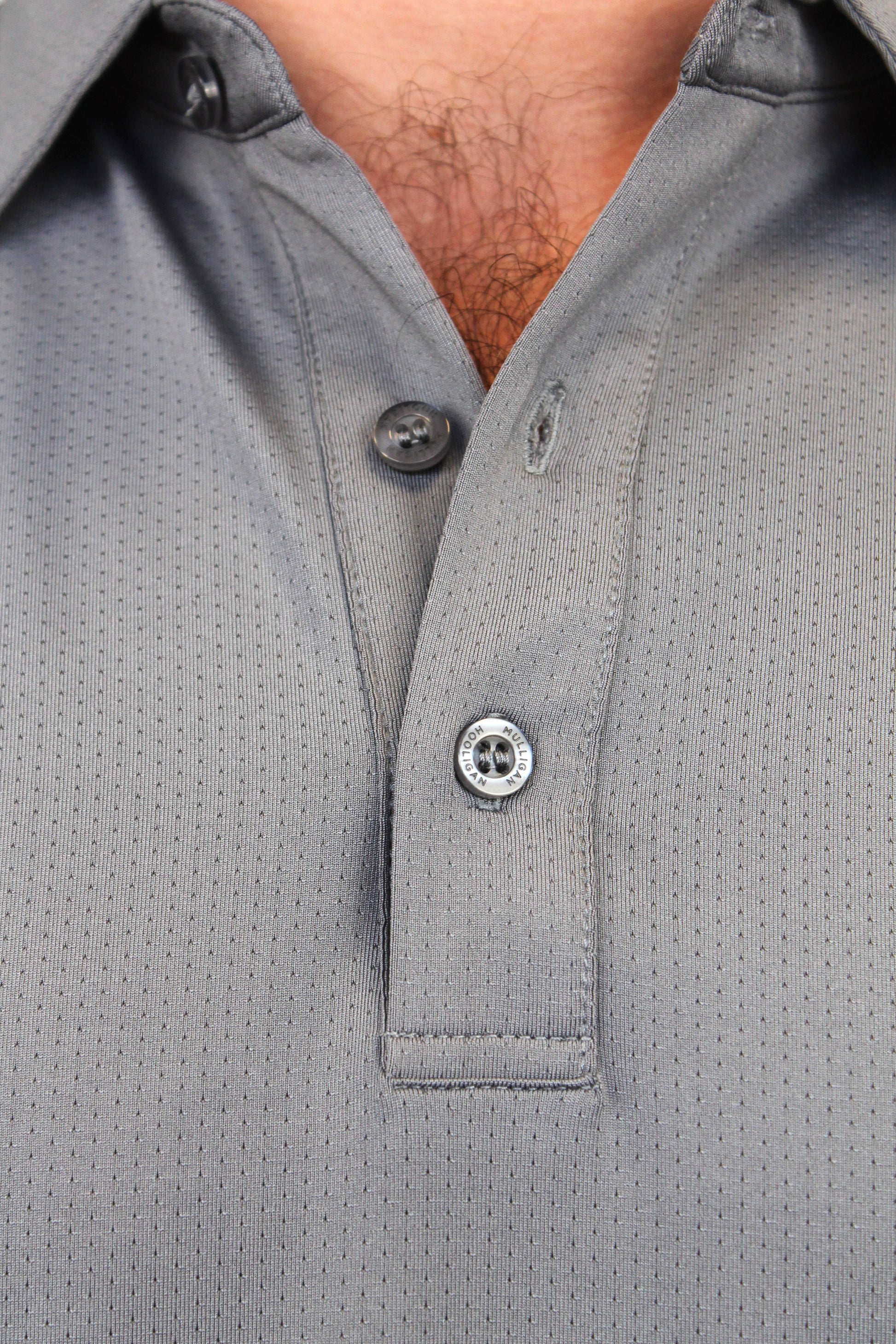 Close-up of a gray short-sleeve polo shirt with a micro-perforated text saying "Mulligan Hooligan', featuring a 3-button placket.