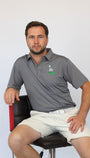 A man sitting on a black and red chair wearing a gray polo shirt with a green and white logo on the left chest, paired with off-white shorts, against a plain light gray background.