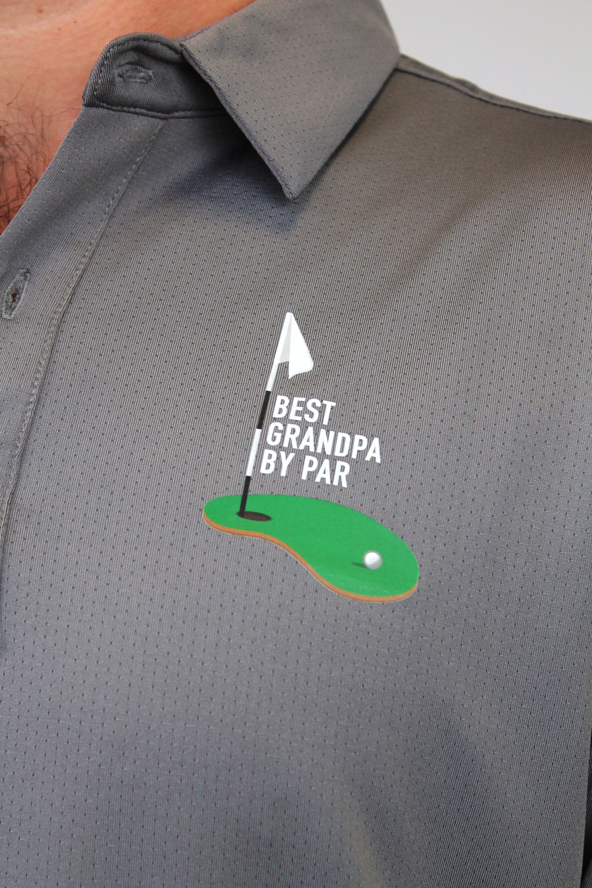 Close-up of a gray perforated polo shirt worn by a person, featuring a golf-themed chest graphic with a green putting green, white flag, and the text 'BEST GRANDPA BY PAR'.