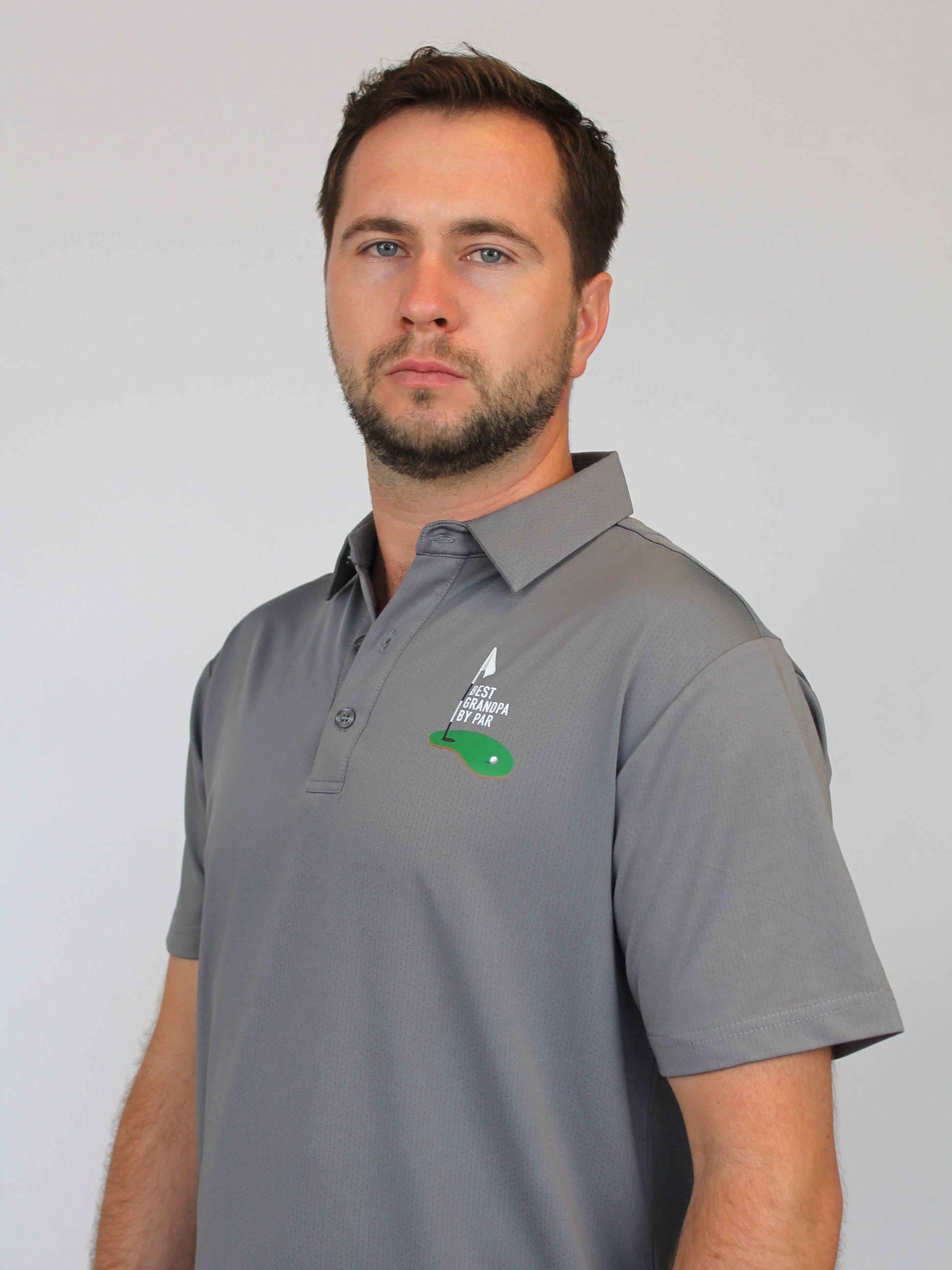 A man wearing a gray short-sleeve polo shirt with a green and white golf logo on the left chest saying 'Best Grandpa by Par', photographed against a plain light gray background.