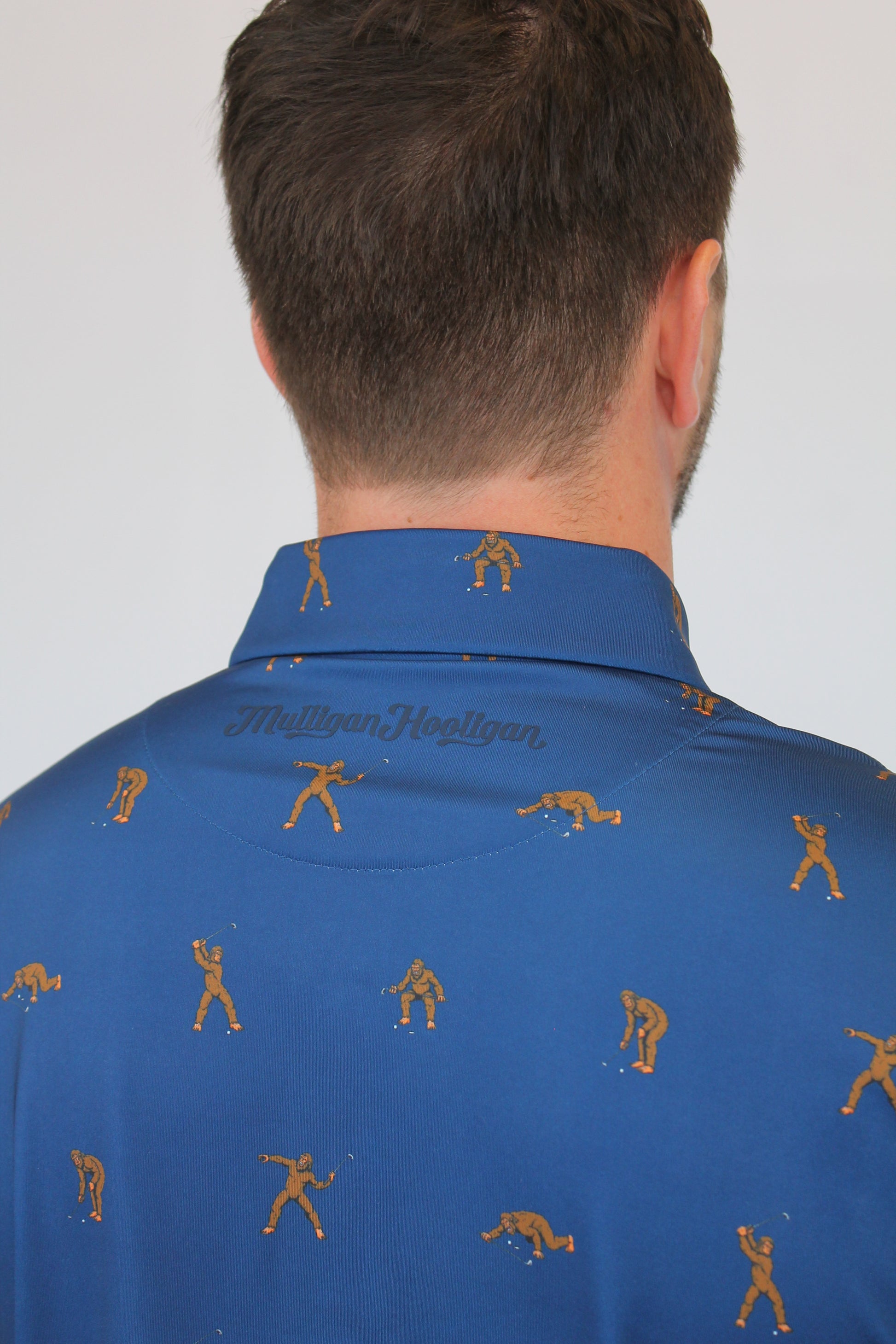 Close-up of a man wearing a blue collared shirt with a repeating brown Golfing Bigfoot figure print, and 'Mulligan Hooligan' Writing on the back, photographed against a plain light background. 