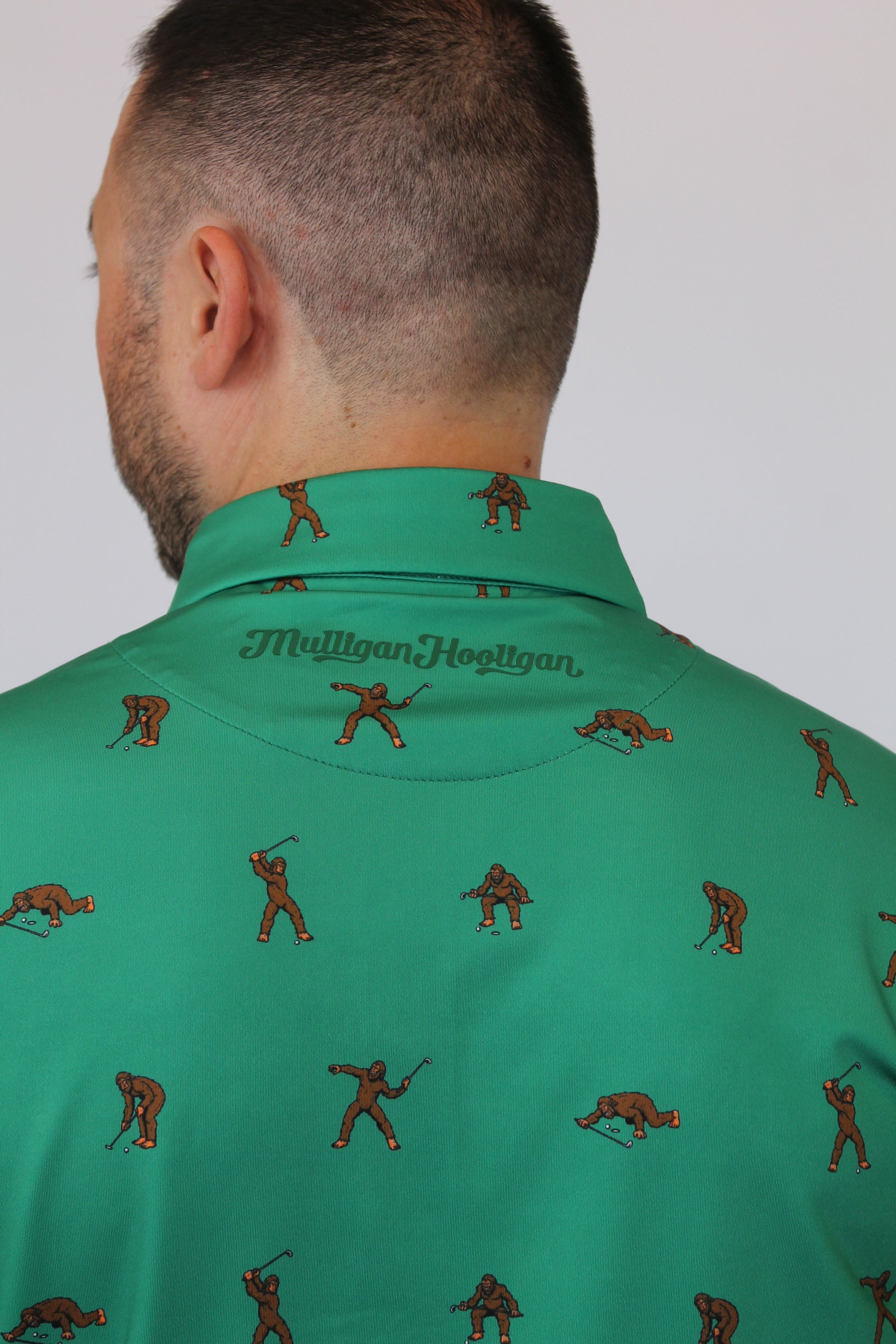 Close-up of a person wearing a green collared shirt with a repeating brown golfers Bigfoot pattern and 'Mulligan Hooligan' text on the collar, against a plain light background.