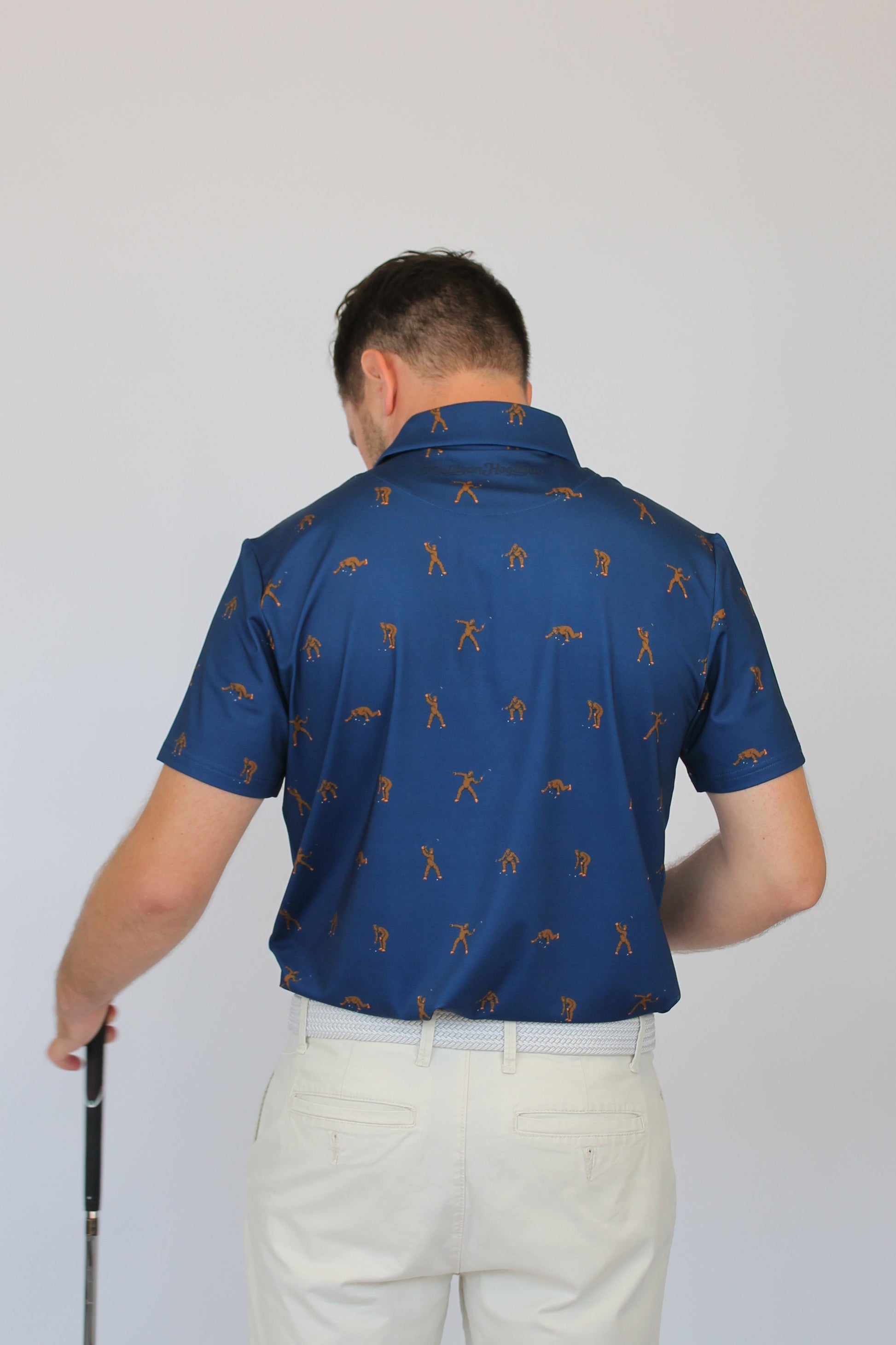 A man viewed from behind wearing a blue short-sleeve polo shirt with a repeating golf player Bigfoot pattern, paired with off-white trousers, holding a golf club against a plain light gray background.