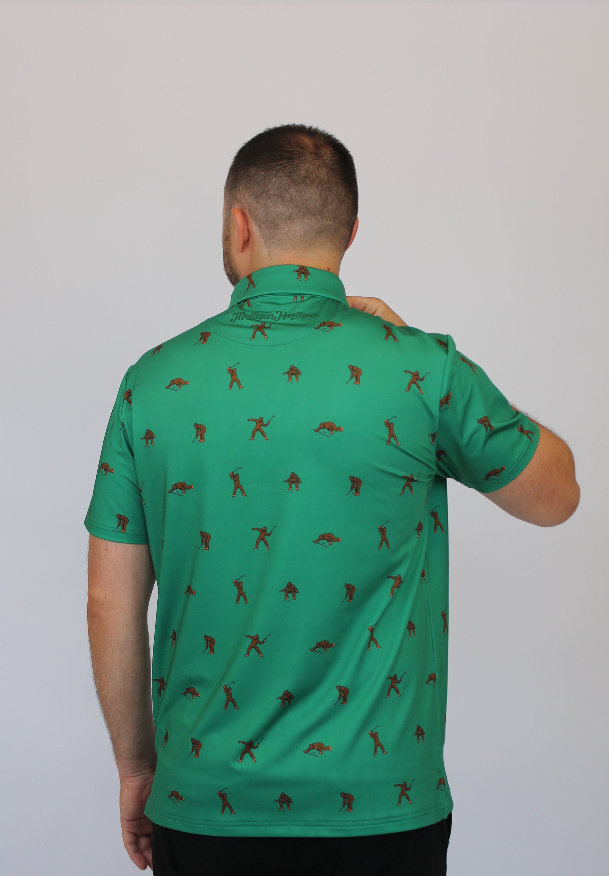 A man viewed from behind wearing the Mulligan Hooligan Bigfoot green short-sleeve polo shirt with a repeating brown golfer silhouette pattern, set against a plain light gray background.