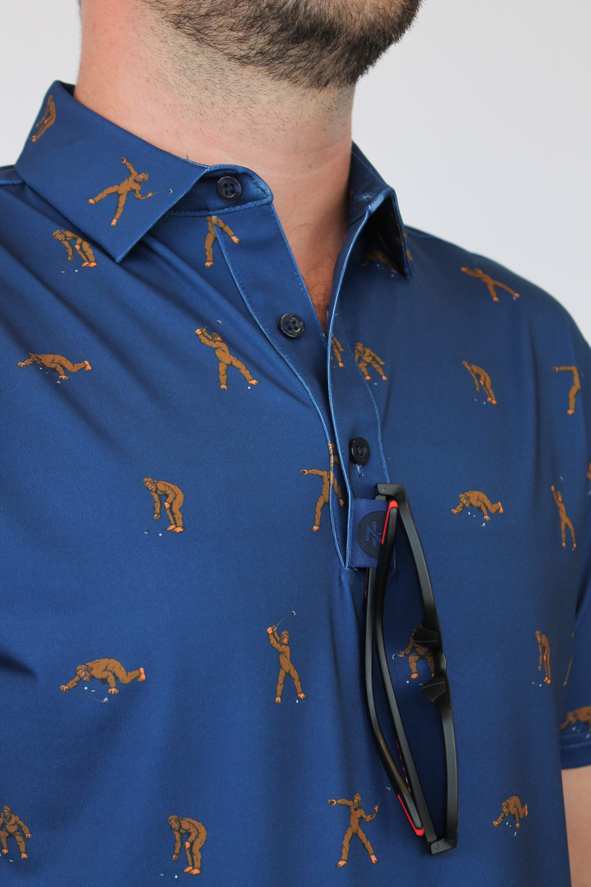 A close-up of a man wearing a blue short-sleeve button-up shirt with Bigfoot golf-themed print of golfers and a golf ball, with black sunglasses hanging from the collar.