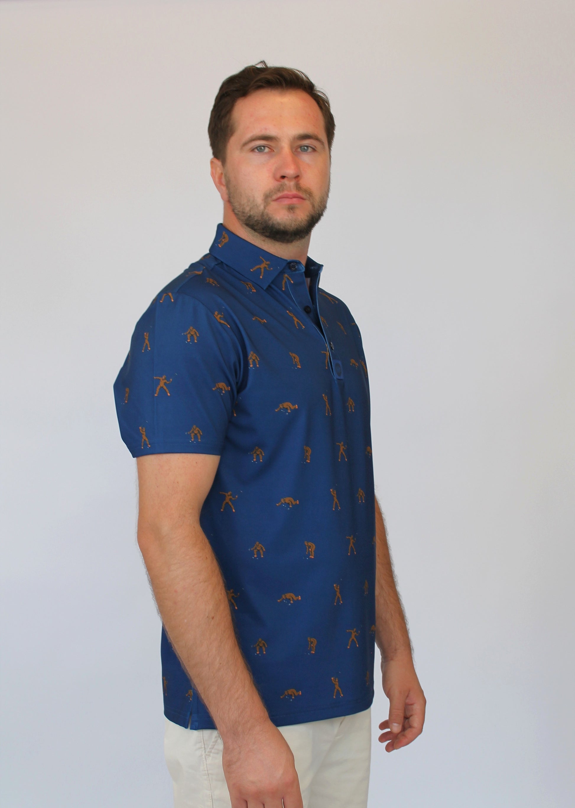 A man wearing a short-sleeve blue polo shirt with small  Golfing Sasquatch golf-themed pattern, paired with off-white trousers, standing against a plain light gray background.