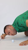 A man in a green Golfing Bigfoot Patterned short-sleeve polo shirt with a brown pattern kneels on a white studio floor, holding a golf club near a white golf ball.