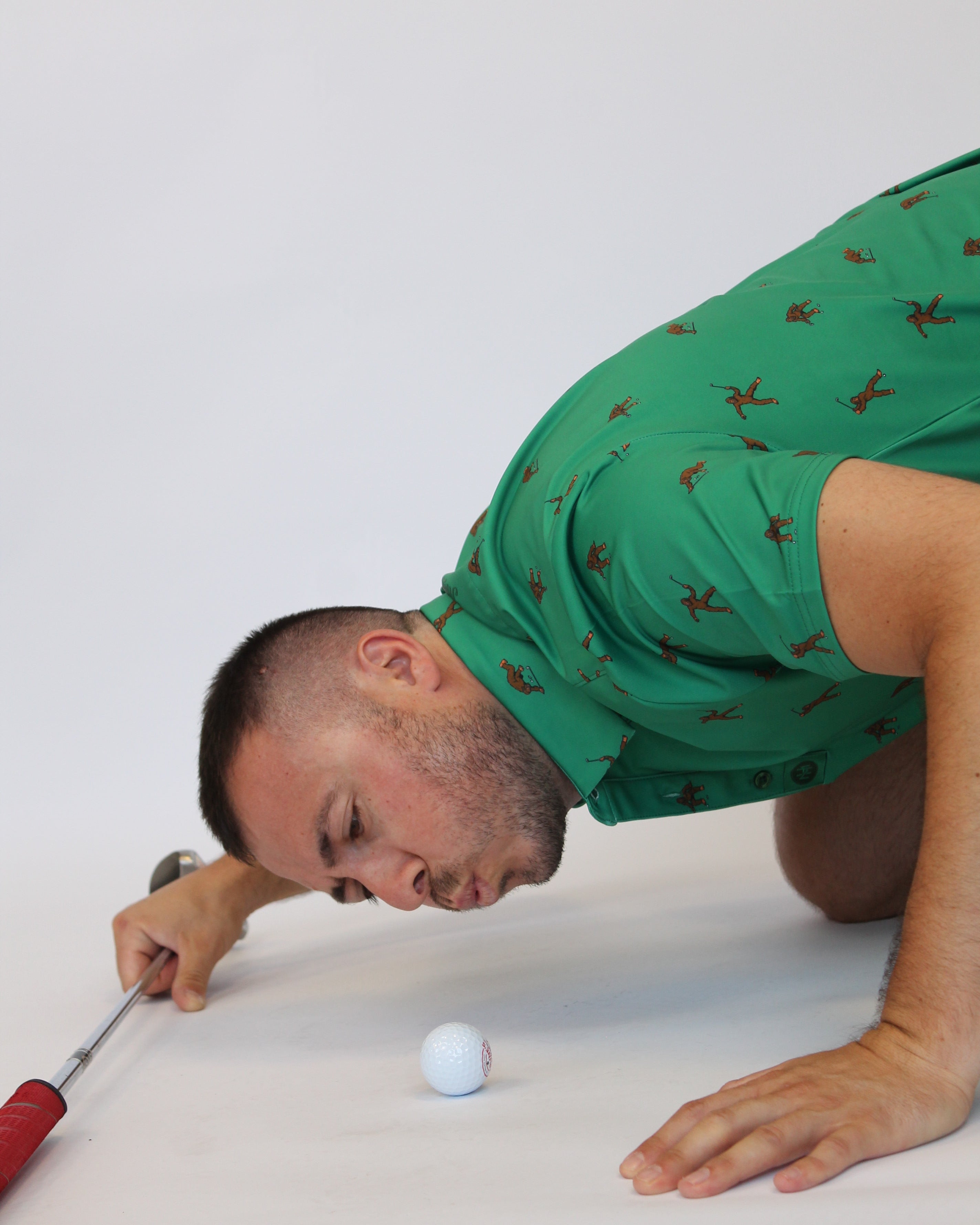 A man in a green Golfing Bigfoot Patterned short-sleeve polo shirt with a brown pattern kneels on a white studio floor, holding a golf club near a white golf ball.
