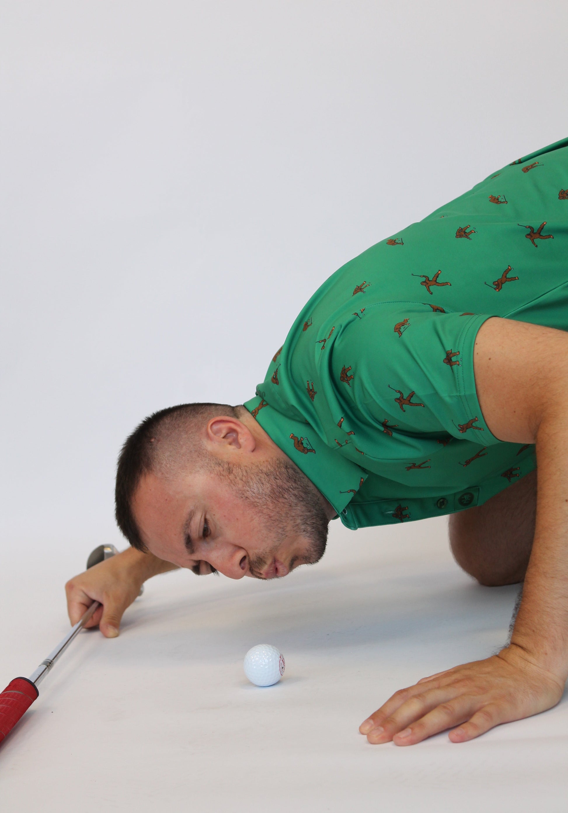 A man in a green Golfing Bigfoot Patterned short-sleeve polo shirt with a brown pattern kneels on a white studio floor, holding a golf club near a white golf ball.