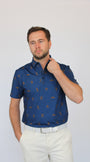 A man wearing a short-sleeve blue polo shirt with a repeating brown golf Bigfoot pattern, paired with off-white trousers, standing against a light gray studio background.