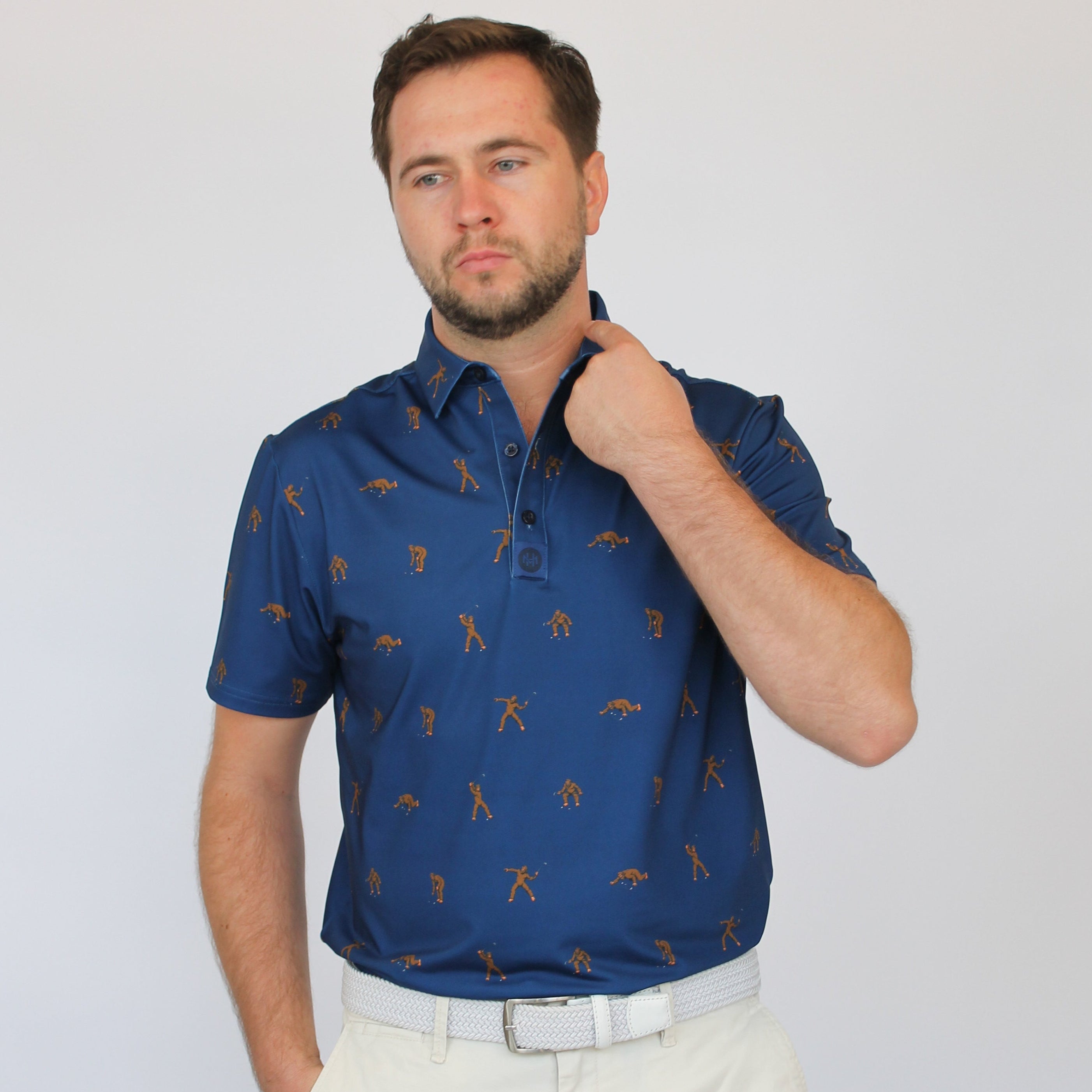 A man wearing a short-sleeve blue polo shirt with a repeating brown golf Bigfoot pattern, paired with off-white trousers, standing against a light gray studio background.