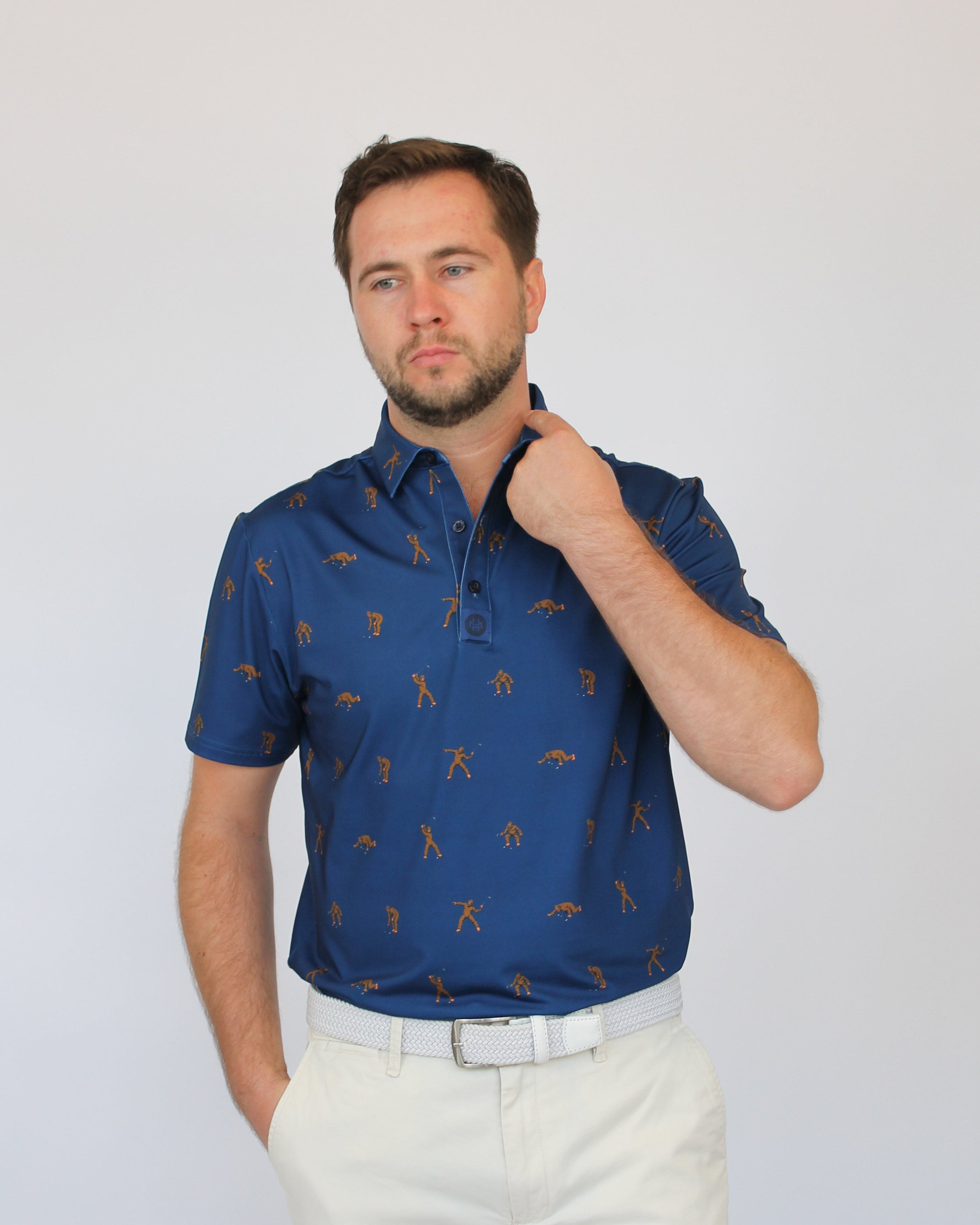 A man wearing a short-sleeve blue polo shirt with a repeating brown golf Bigfoot pattern, paired with off-white trousers, standing against a light gray studio background.