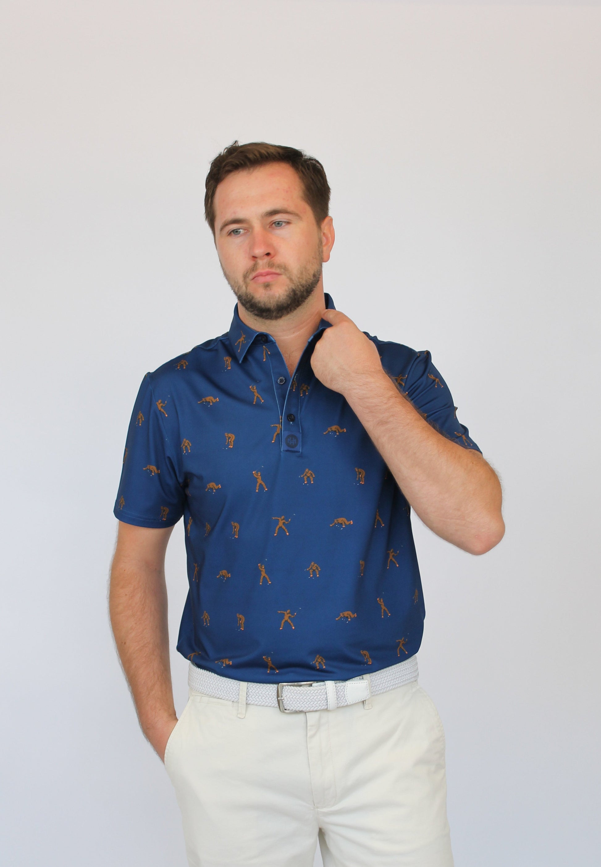 A man wearing a short-sleeve blue polo shirt with a repeating brown golf Bigfoot pattern, paired with off-white trousers, standing against a light gray studio background.