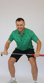 A man in a green Golfing Bigfoot patterned polo shirt and black shorts crouches while holding a golf club horizontally in front of him, against a plain light gray studio background.