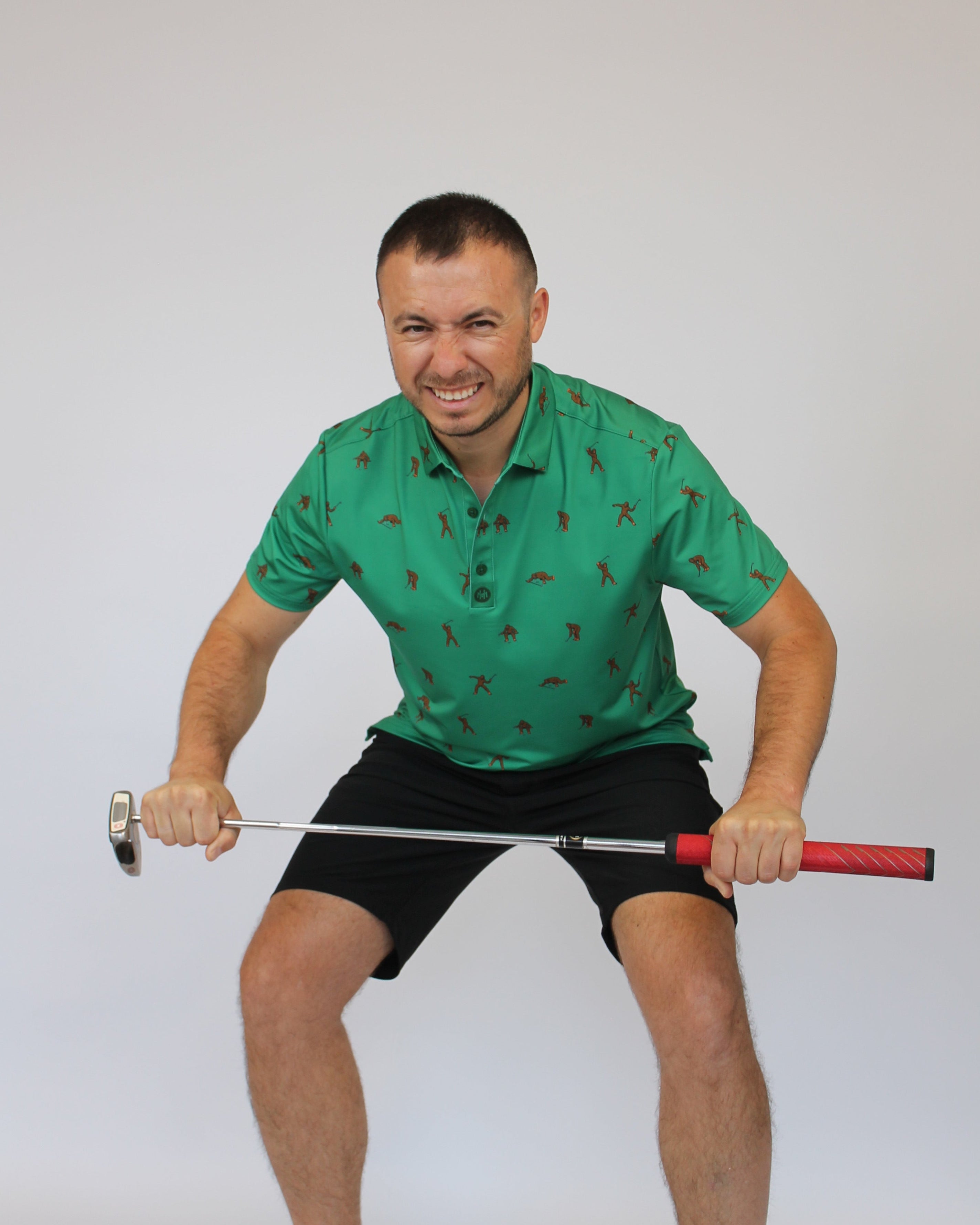 A man in a green Golfing Bigfoot patterned polo shirt and black shorts crouches while holding a golf club horizontally in front of him, against a plain light gray studio background.