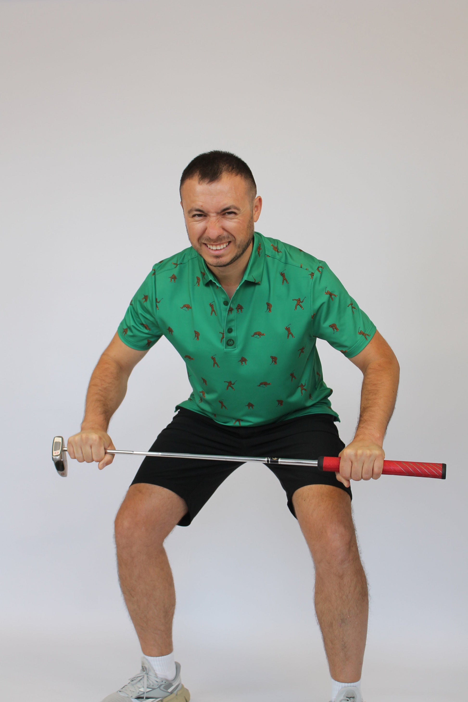 A man in a green Golfing Bigfoot patterned polo shirt and black shorts crouches while holding a golf club horizontally in front of him, against a plain light gray studio background.