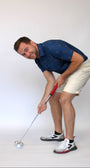 A man in a blue Golfing Bigfoot patterned short-sleeve shirt and beige shorts is bent over, holding a golf club with a red grip, about to hit a white golf ball on a light gray studio background.