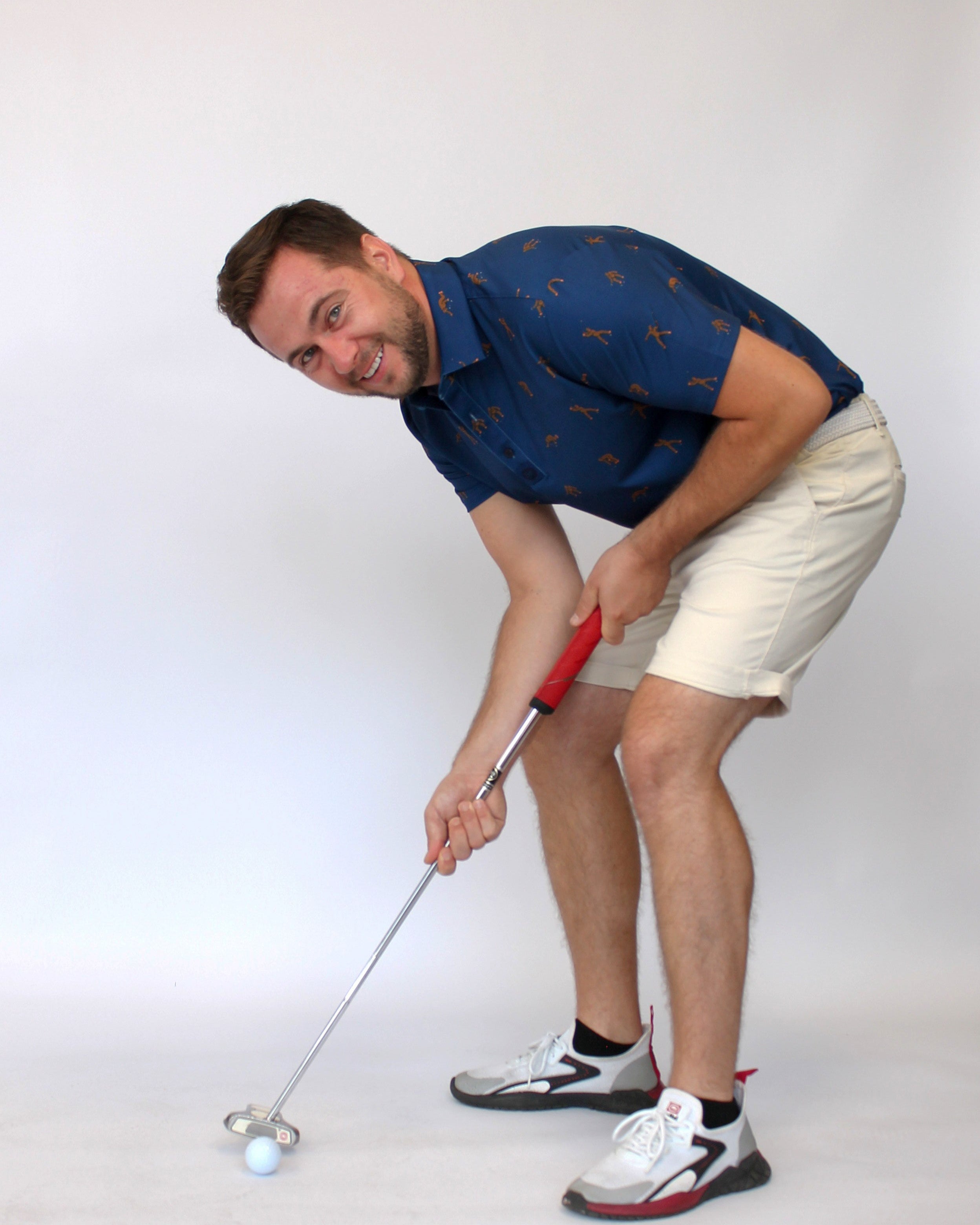 A man in a blue Golfing Bigfoot patterned short-sleeve shirt and beige shorts is bent over, holding a golf club with a red grip, about to hit a white golf ball on a light gray studio background.