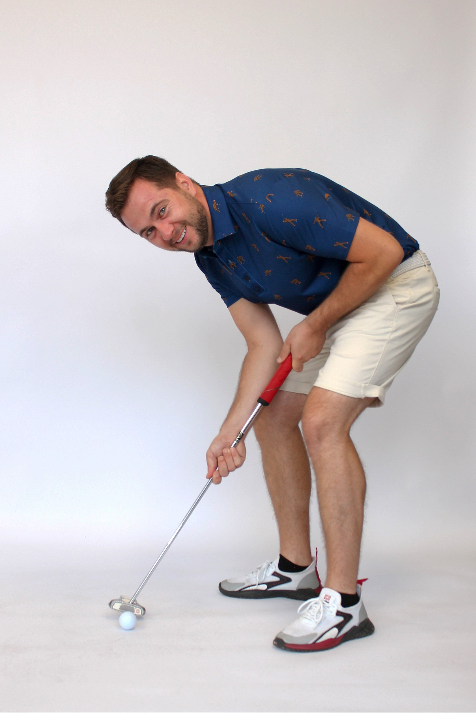 A man in a blue Golfing Bigfoot patterned short-sleeve shirt and beige shorts is bent over, holding a golf club with a red grip, about to hit a white golf ball on a light gray studio background.