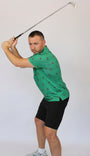 A man in a green Bigfoot patterned polo shirt and black shorts is captured mid-swing with a golf club against a plain light gray studio background.