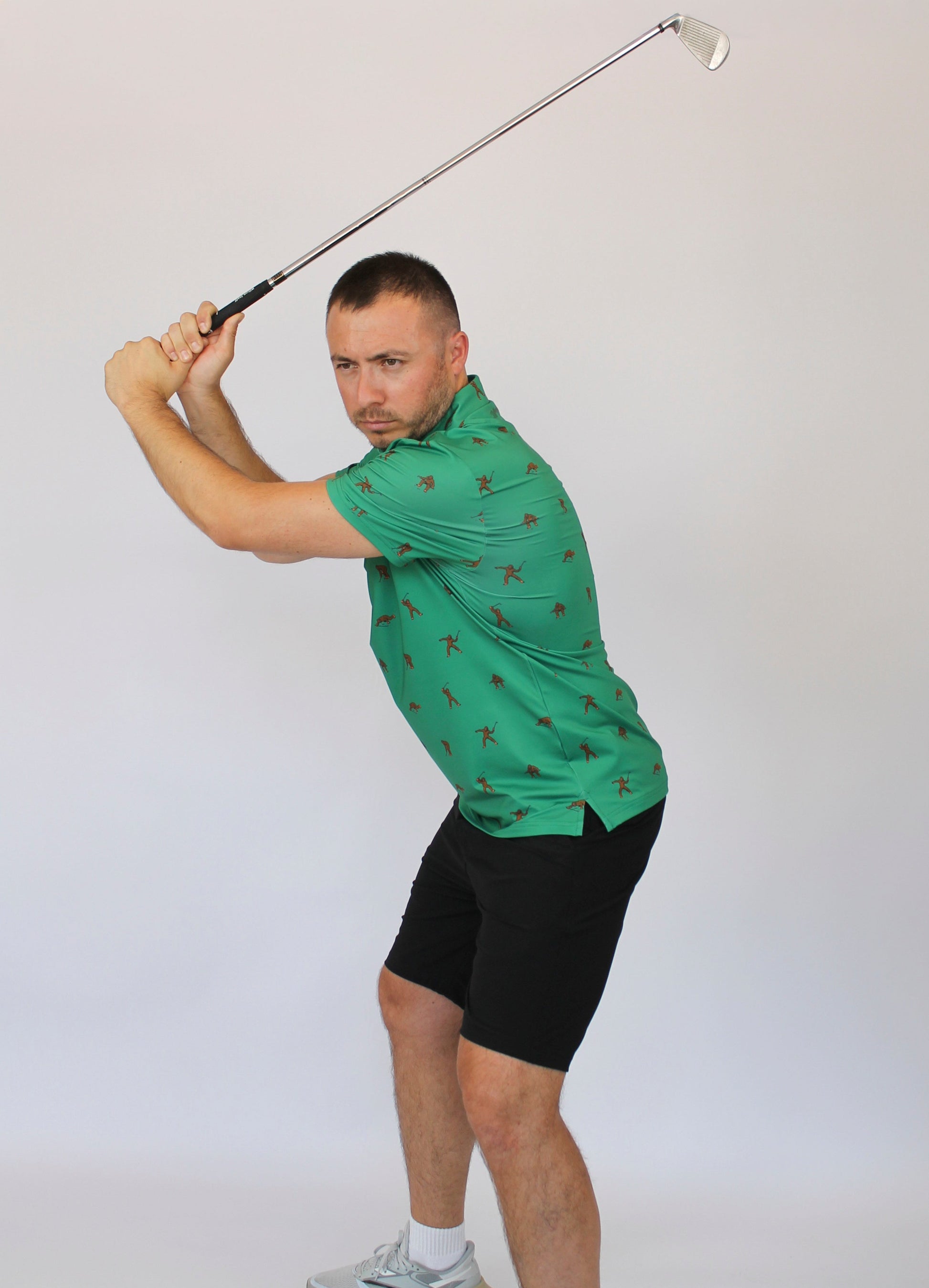A man in a green Bigfoot patterned polo shirt and black shorts is captured mid-swing with a golf club against a plain light gray studio background.