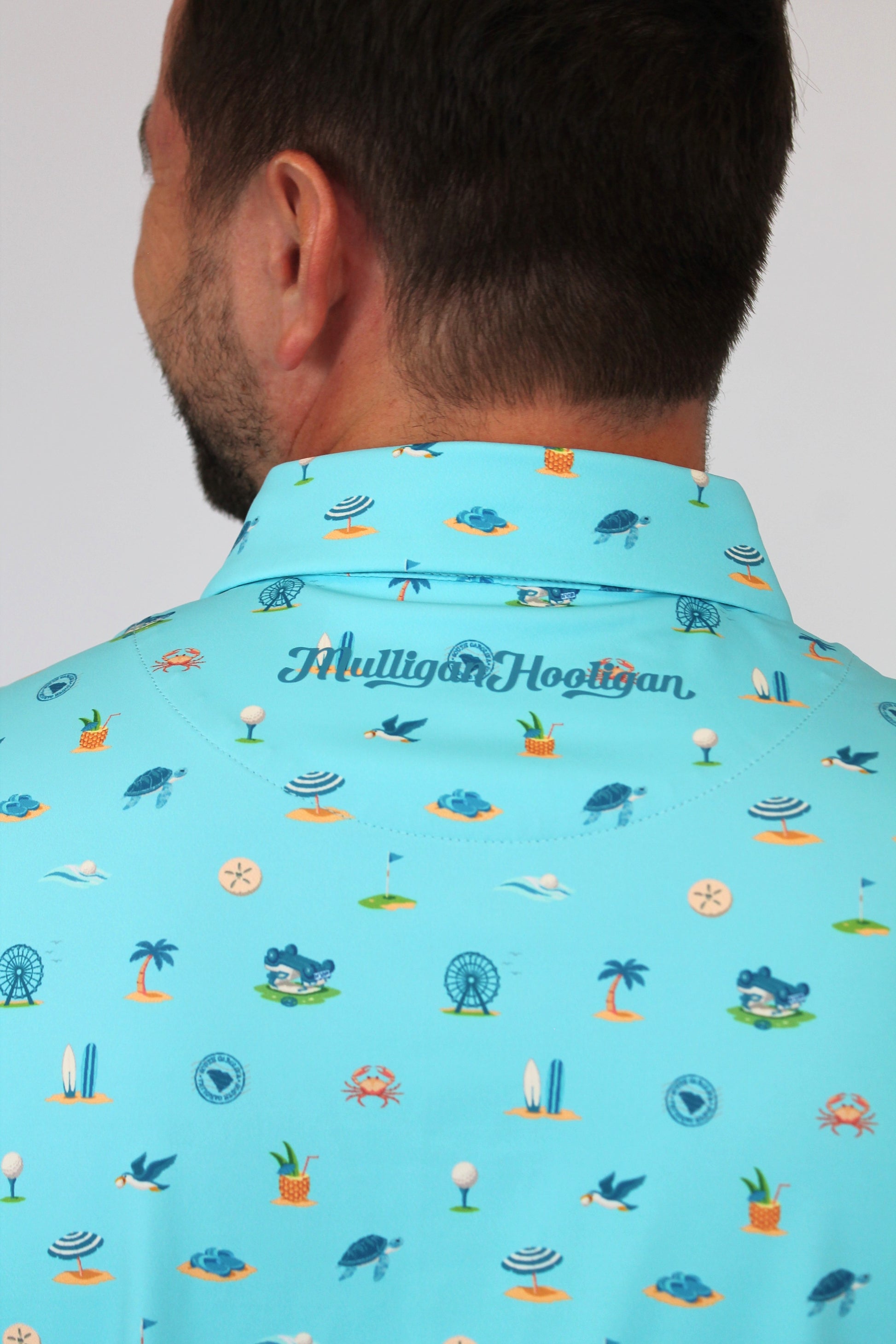 A man shown from the back wearing a light blue collared shirt with a colorful golf-themed and beach-theme print of tees, greens, holes, and golf carts. The brand name 'Mulligan Hooligan' is printed in script across the collar.
