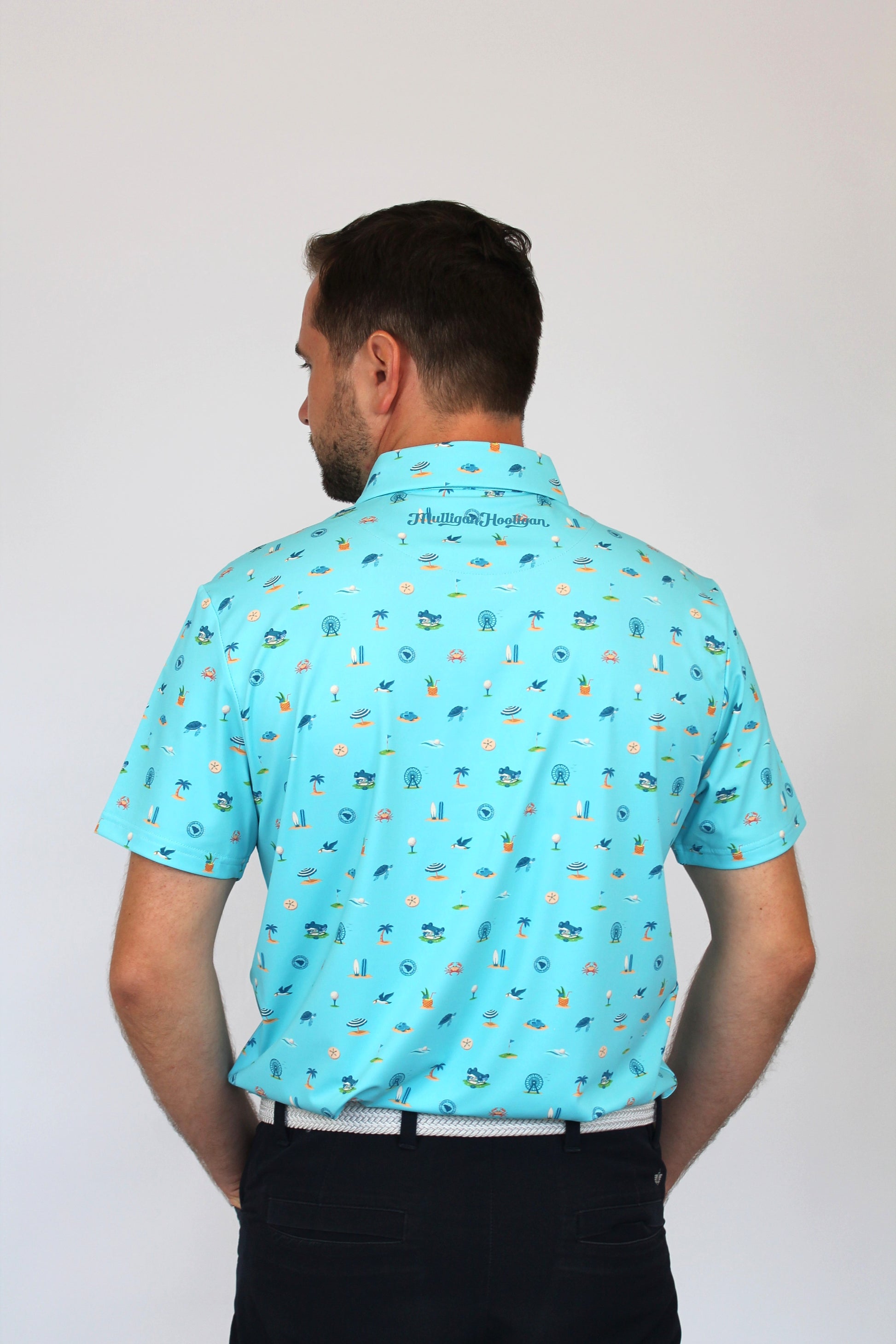 A man viewed from behind wearing a light blue short-sleeve collared shirt with a small all-over Coastal Links, Myrtle Beach, SC, Beach-theme pattern, paired with black pants, against a plain light gray background.