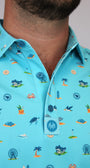 A close-up of a man wearing a light blue short-sleeve button-up shirt with a colorful beach-themed print Myrtle Beach, South Carolina, including palm trees, crabs, seashells, and birds. The shirt features a collar and a button placket with a circular 'MH' logo patch near the bottom button.