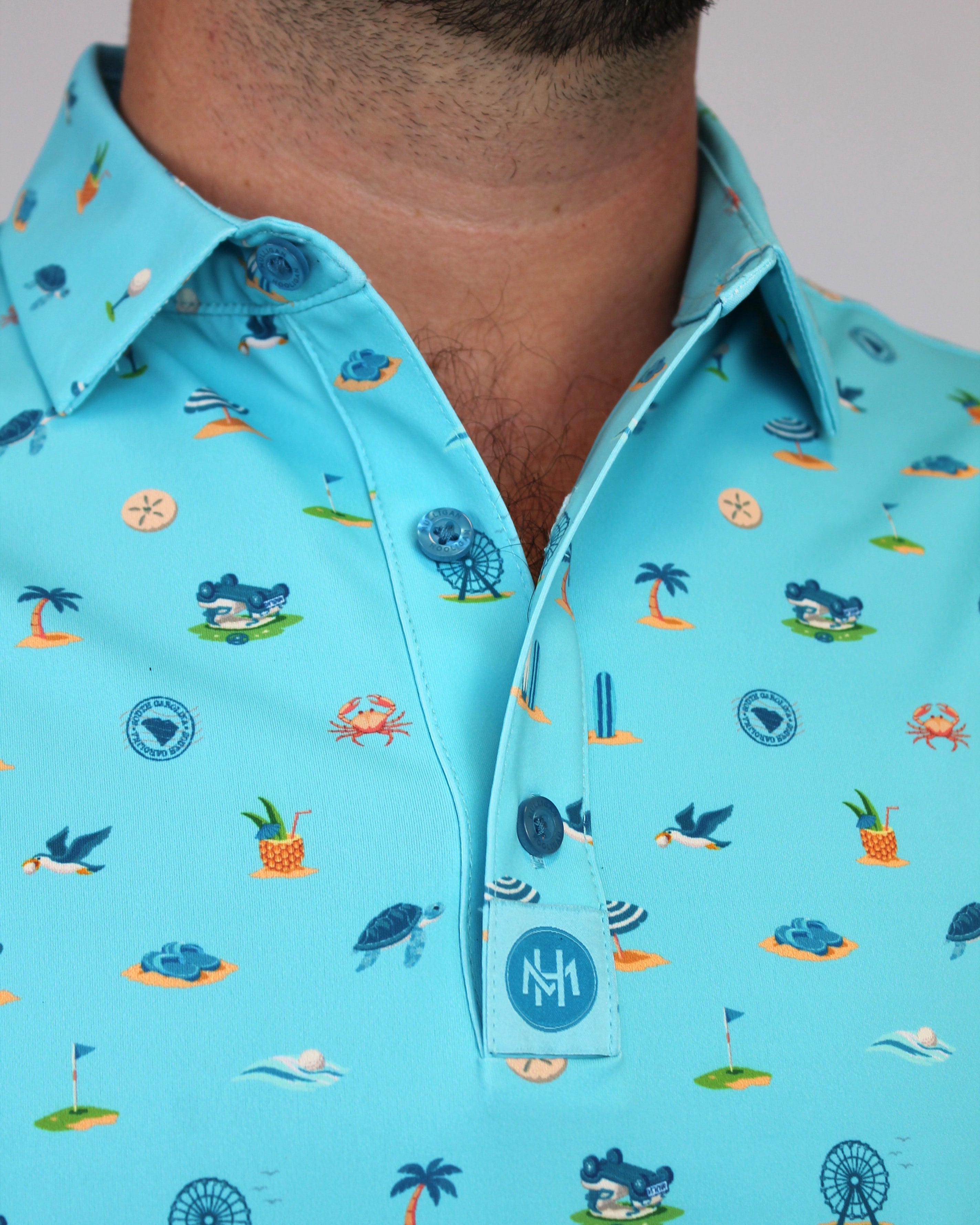 A close-up of a man wearing a light blue short-sleeve button-up shirt with a colorful beach-themed print Myrtle Beach, South Carolina, including palm trees, crabs, seashells, and birds. The shirt features a collar and a button placket with a circular 'MH' logo patch near the bottom button.