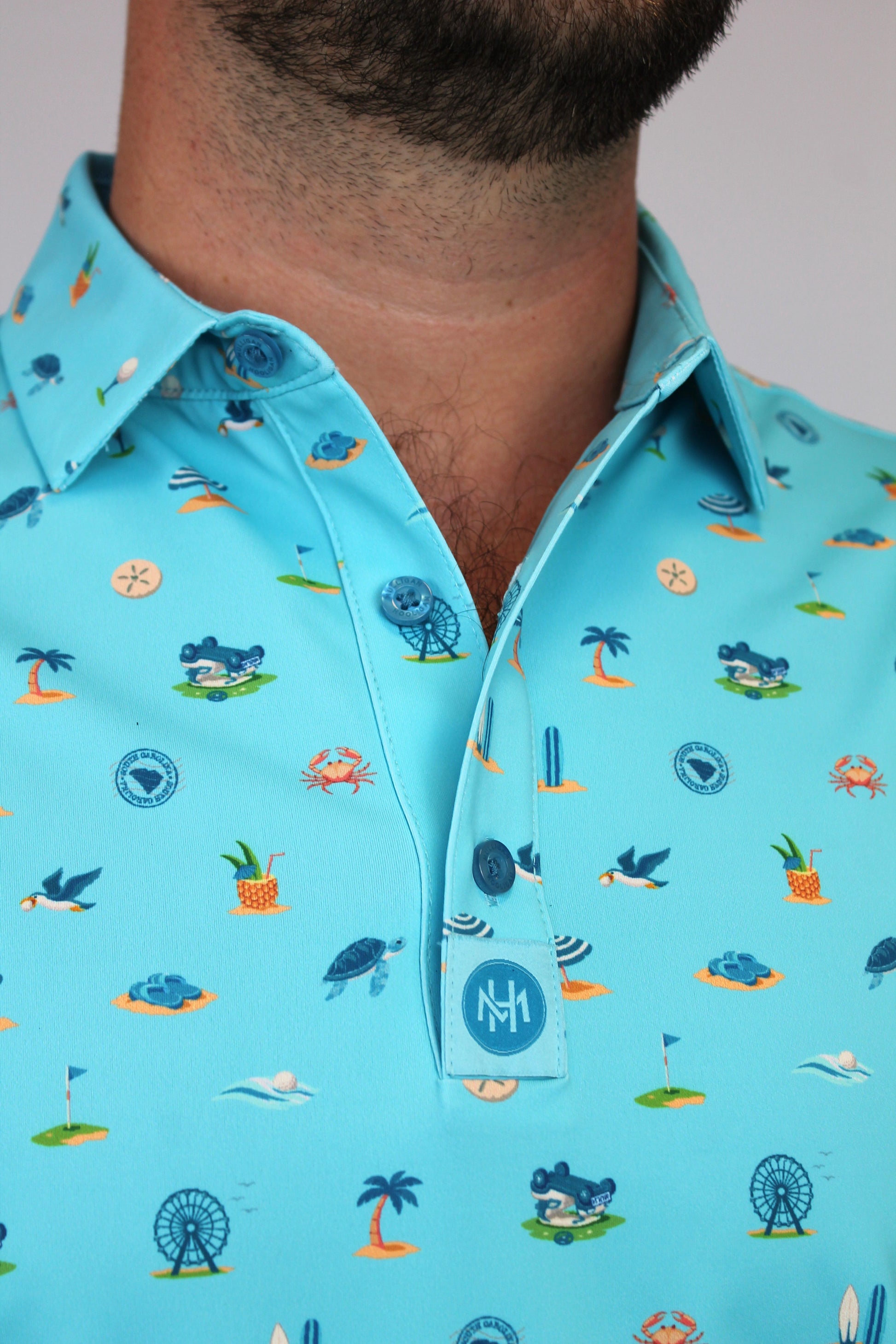 A close-up of a man wearing a light blue short-sleeve button-up shirt with a colorful beach-themed print Myrtle Beach, South Carolina, including palm trees, crabs, seashells, and birds. The shirt features a collar and a button placket with a circular 'MH' logo patch near the bottom button.