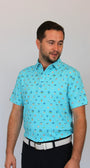 A man wearing a light blue short-sleeve polo shirt with a small beach-theme, Myrtle Beach, South Carolina all-over pattern, paired with a white belt and dark trousers, standing against a plain light gray background.