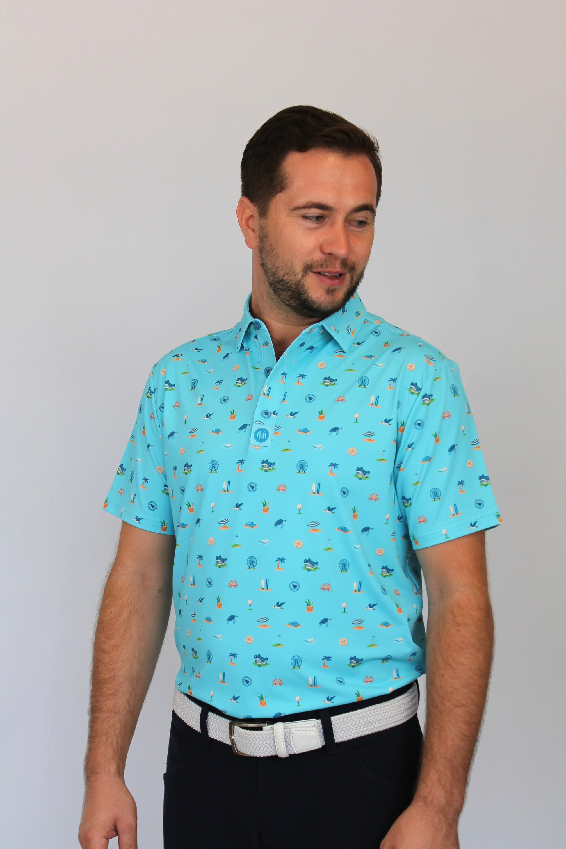 A man wearing a light blue short-sleeve polo shirt with a small beach-theme, Myrtle Beach, South Carolina all-over pattern, paired with a white belt and dark trousers, standing against a plain light gray background.