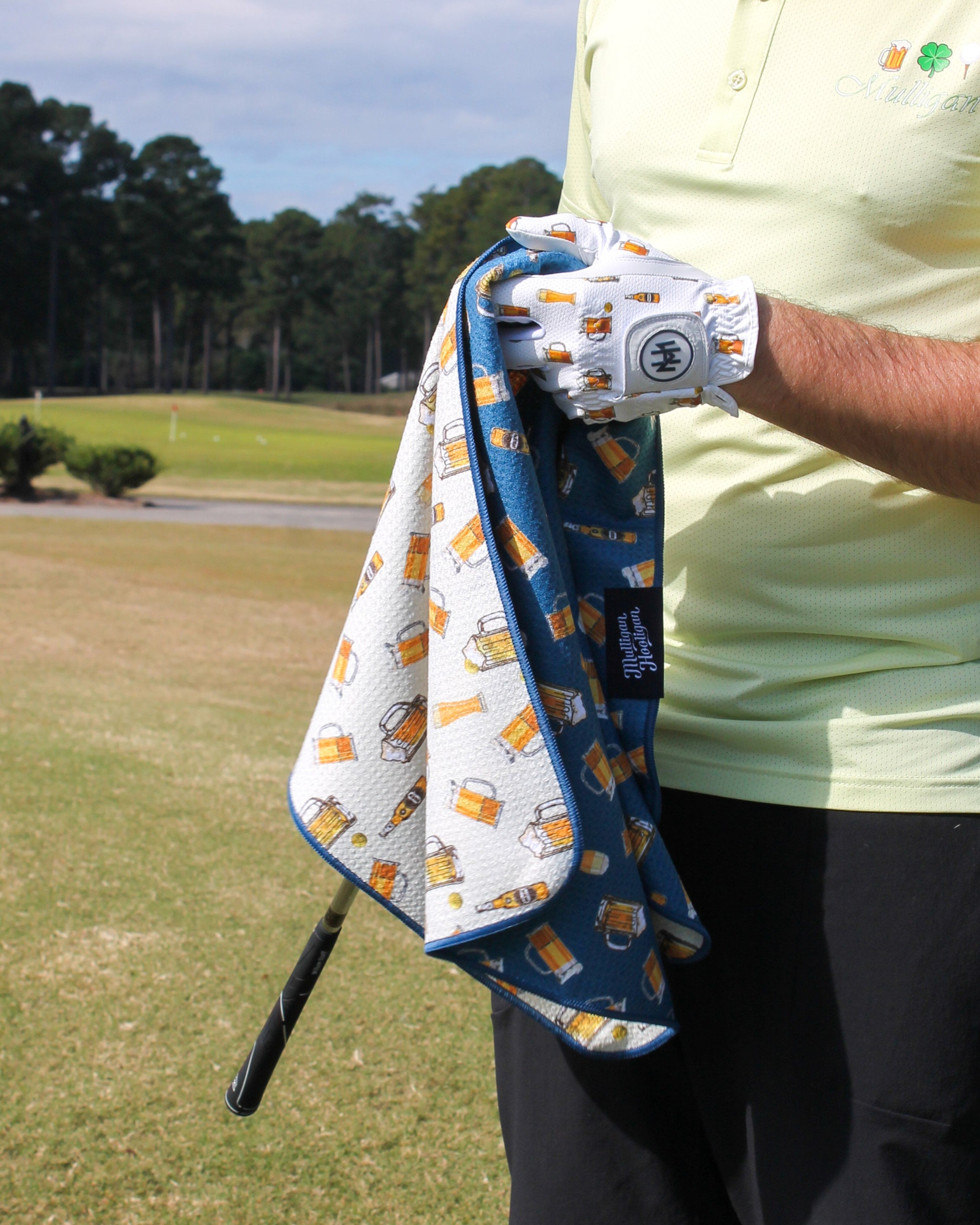 A man on a golf course wearing a light green polo shirt and black pants, holding a patterned golf towel and a golf club. The towel has a white base with a blue border and a repeating print of beer glasses and a golf ball.