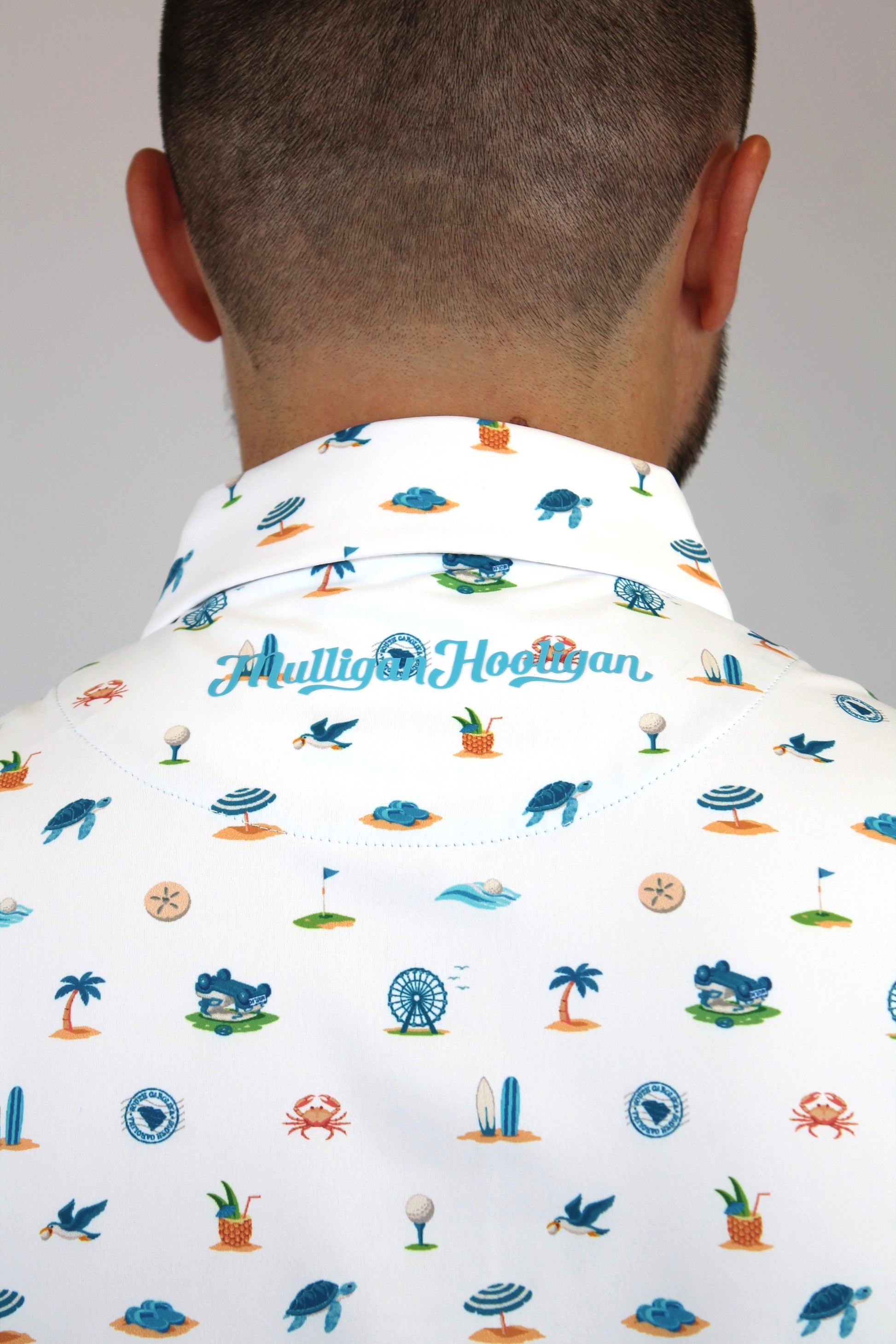 Back view of a man wearing a white collared shirt with a colorful 'Mulligan Hooligan' golf-themed print, featuring blue, green, and orange illustrations of golf course elements.
