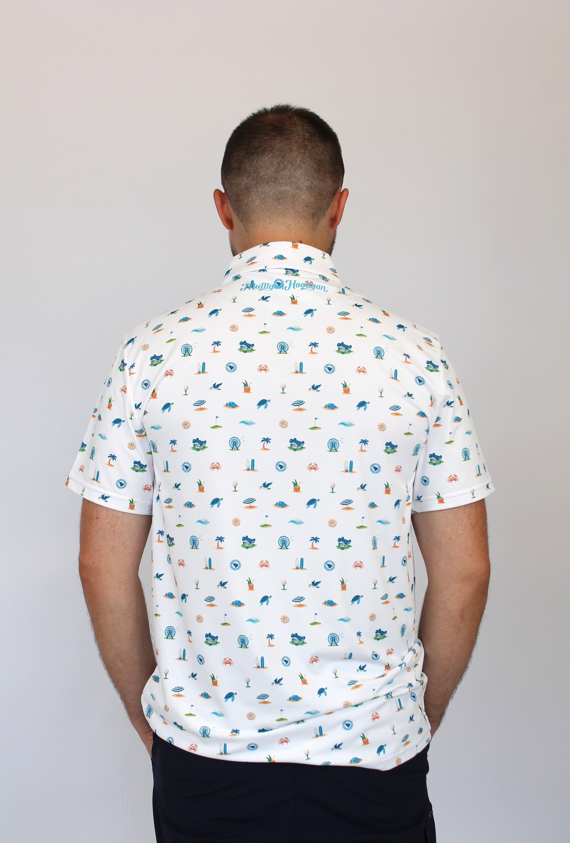 A man viewed from behind wearing a white short-sleeve button-up shirt with a colorful beach-themed print of golf carts, golf flag, SkyWheel, beach, and sea creatures, paired with dark pants, against a plain light gray background.