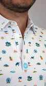 Close-up of a man wearing a white short-sleeve button-up shirt with a colorful beach-themed & golf-theme print including palm trees, crabs, seashells, and waves. A small blue circular logo patch is visible on the pocket.