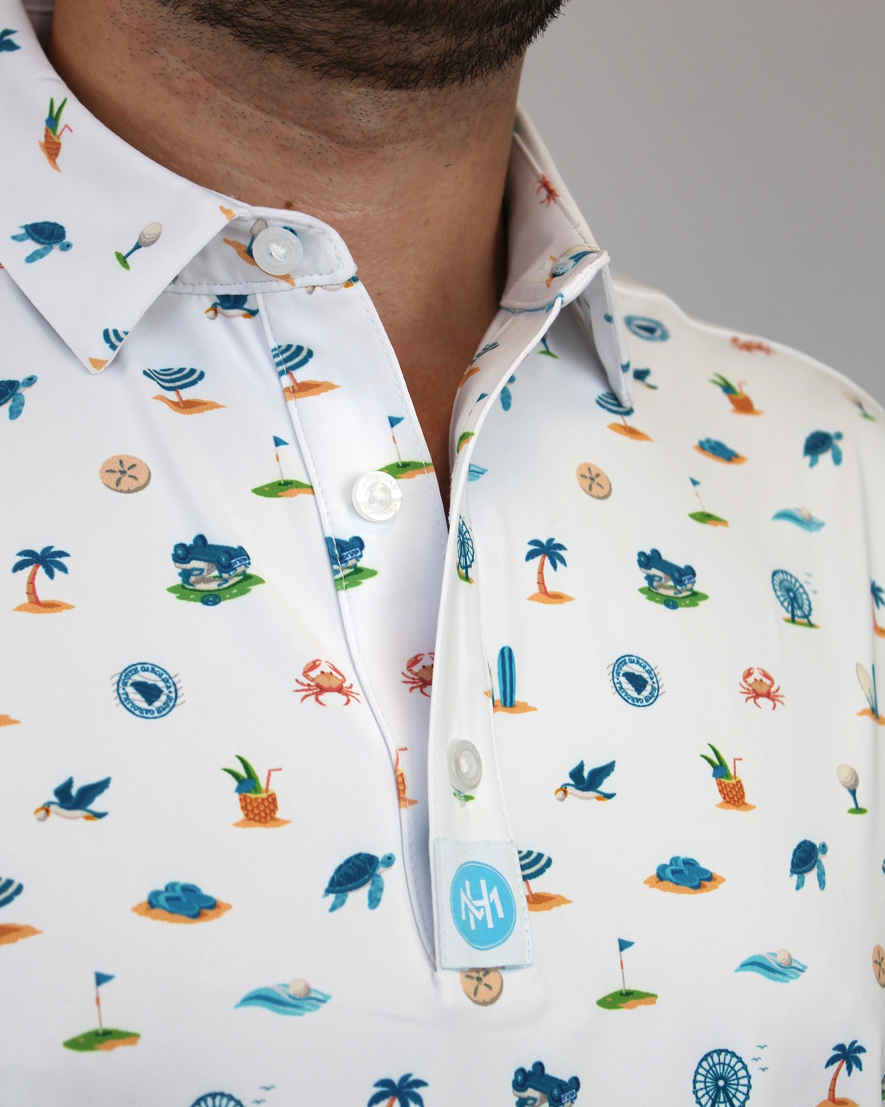 Close-up of a man wearing a white short-sleeve button-up shirt with a colorful beach-themed & golf-theme print including palm trees, crabs, seashells, and waves. A small blue circular logo patch is visible on the pocket.