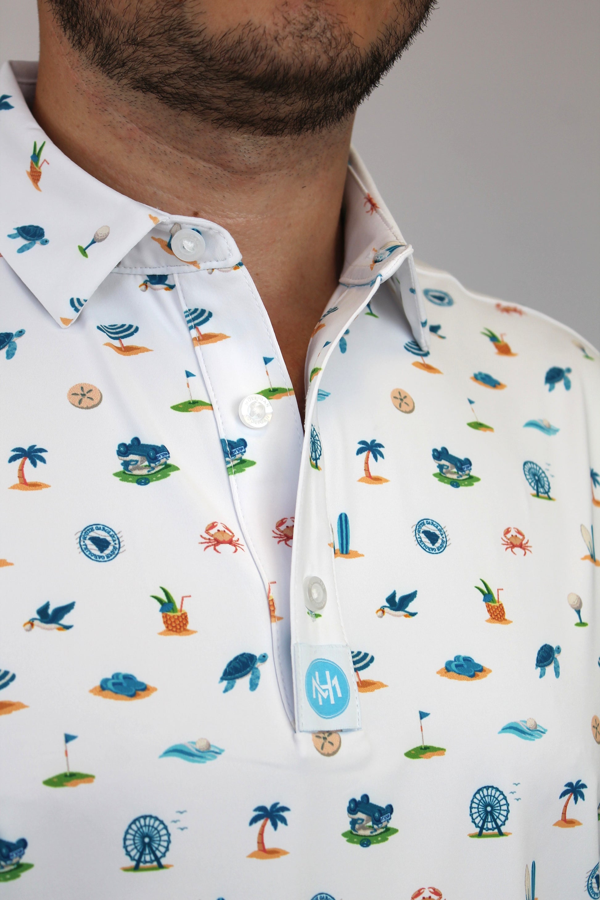 Close-up of a man wearing a white short-sleeve button-up shirt with a colorful beach-themed & golf-theme print including palm trees, crabs, seashells, and waves. A small blue circular logo patch is visible on the pocket.