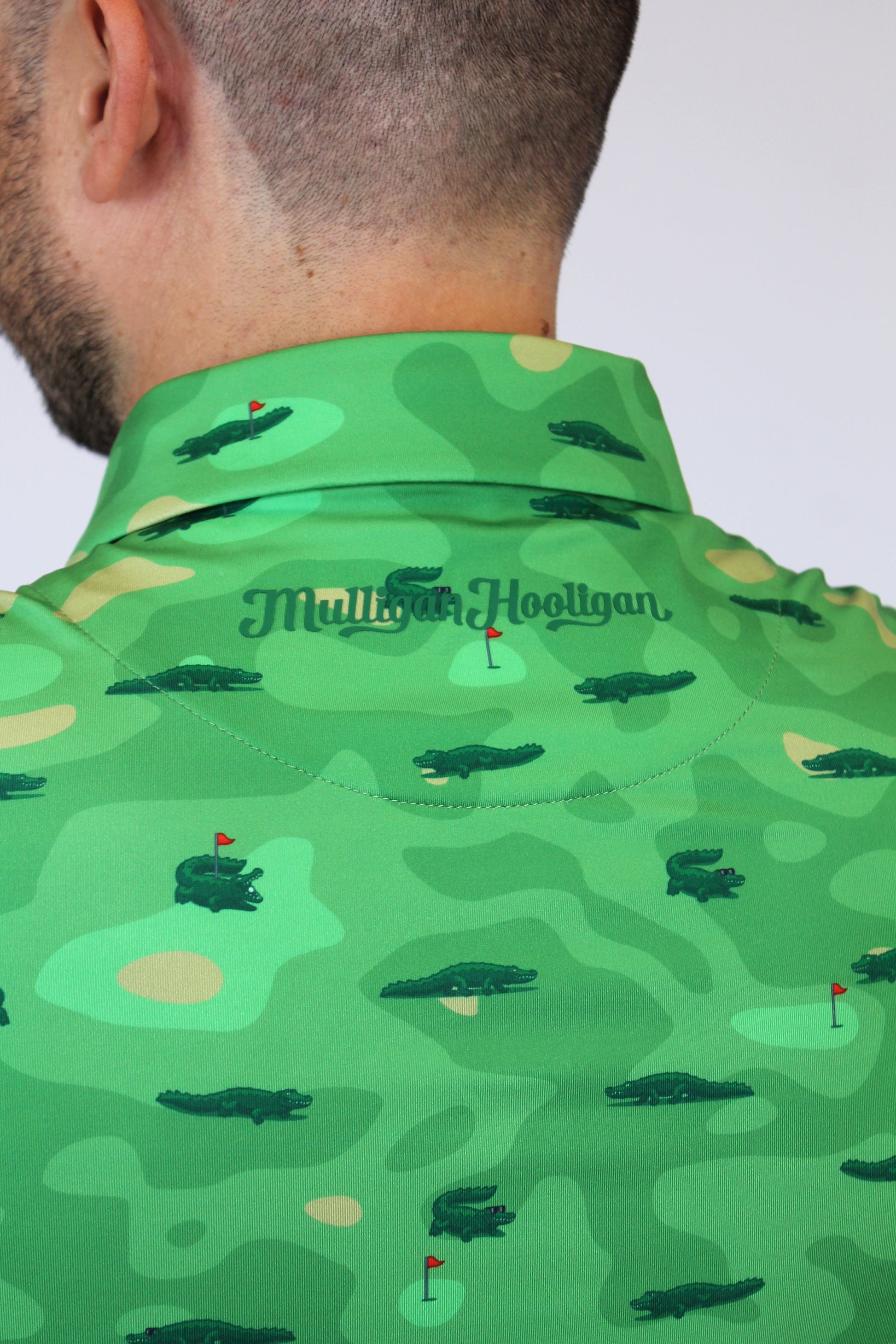 Close-up back view of a man wearing a green collared shirt with an alligator camouflage pattern and the 'Mulligan Hooligan' logo on the collar.