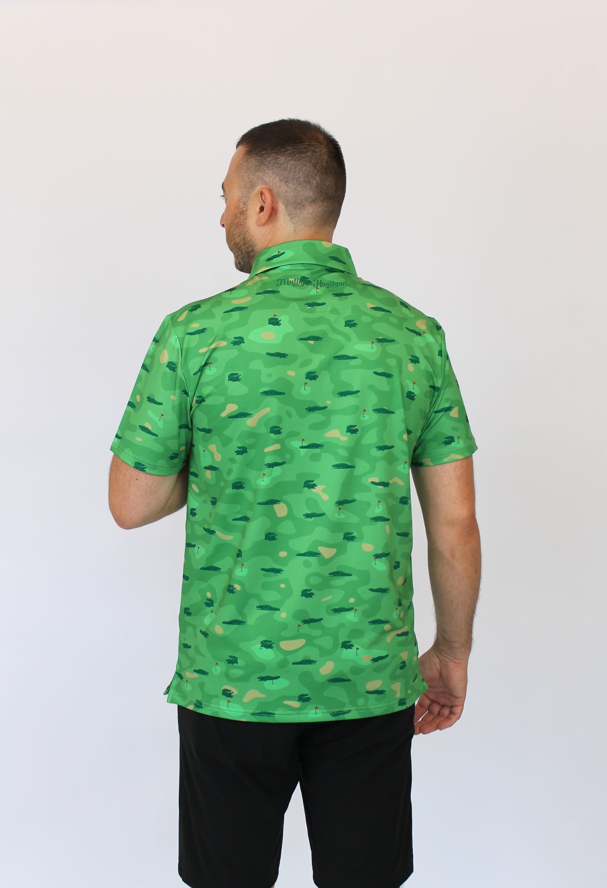 A man viewed from behind wearing a camo green short-sleeve button-up shirt with alligators on a golf coarse print in dark green, paired with black shorts, against a plain white background.