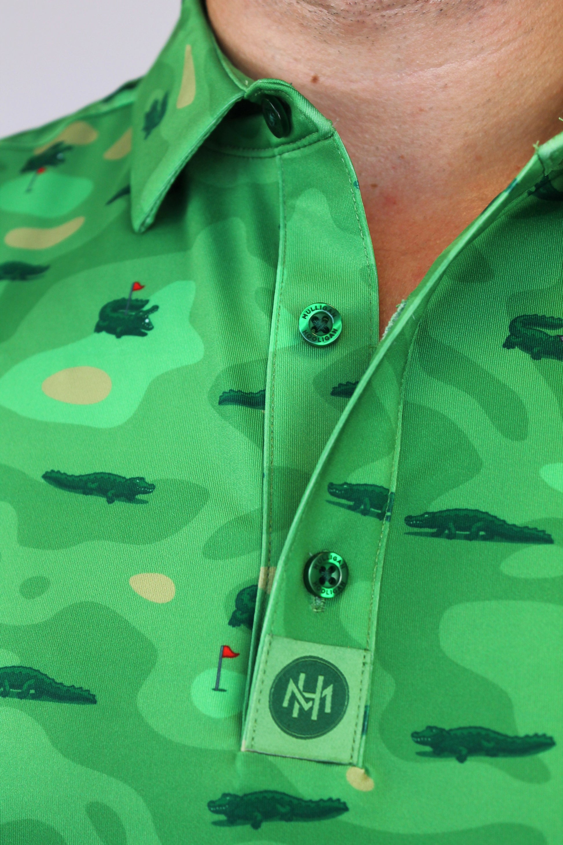 Close-up of a green collared button-up shirt with an alligator and golf course pattern, featuring green buttons and a branded 'Mulligan Hooligan' Custom Engrave.