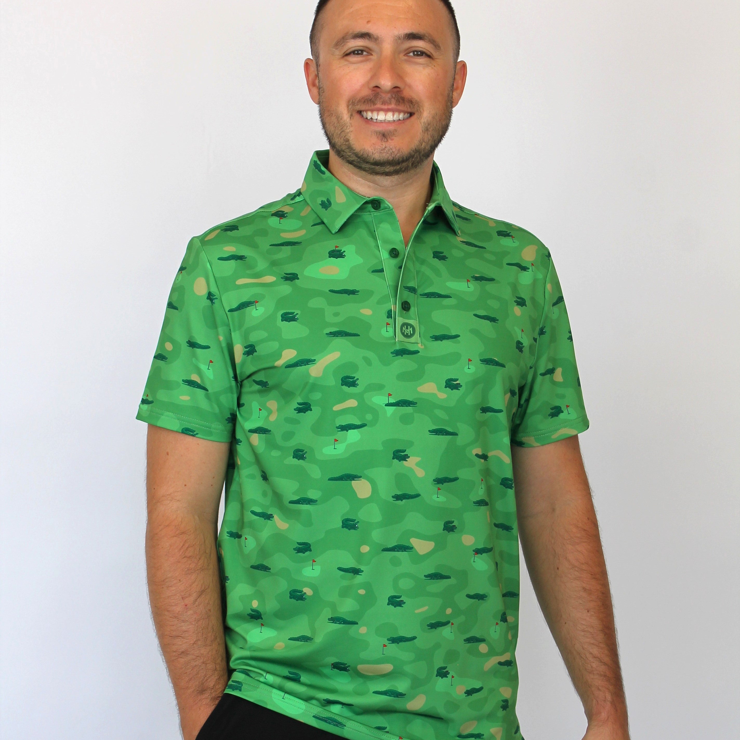 A man wearing a green short-sleeve polo shirt with an all-over camouflage pattern and Alligators on a golf course some with sunglasses, some with a golf ball in their mouth, paired with black pants, standing against a plain white background.